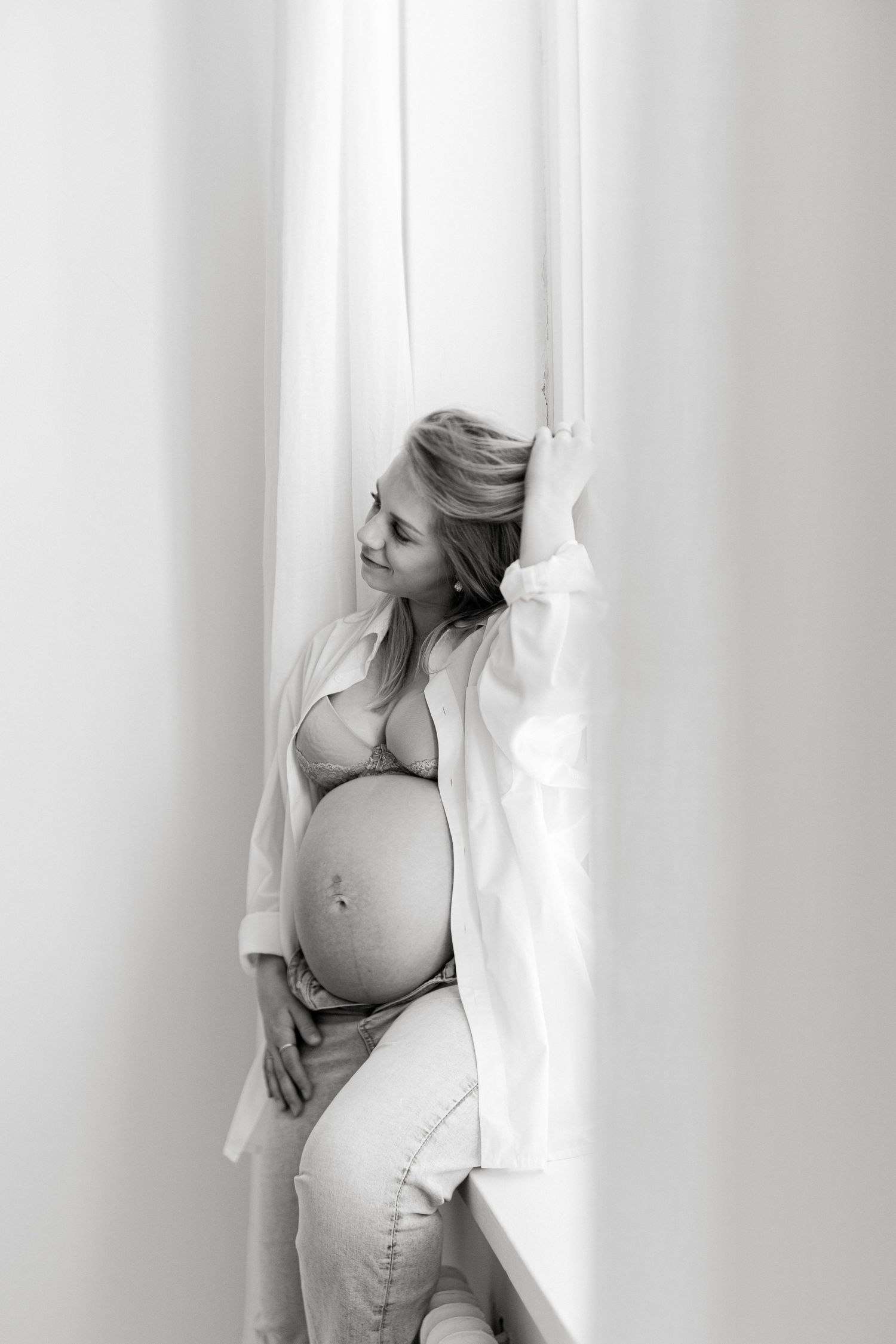 Black and white photo of a pregnant woman in lingerie and open white shirt posing by sheer curtains near a window.