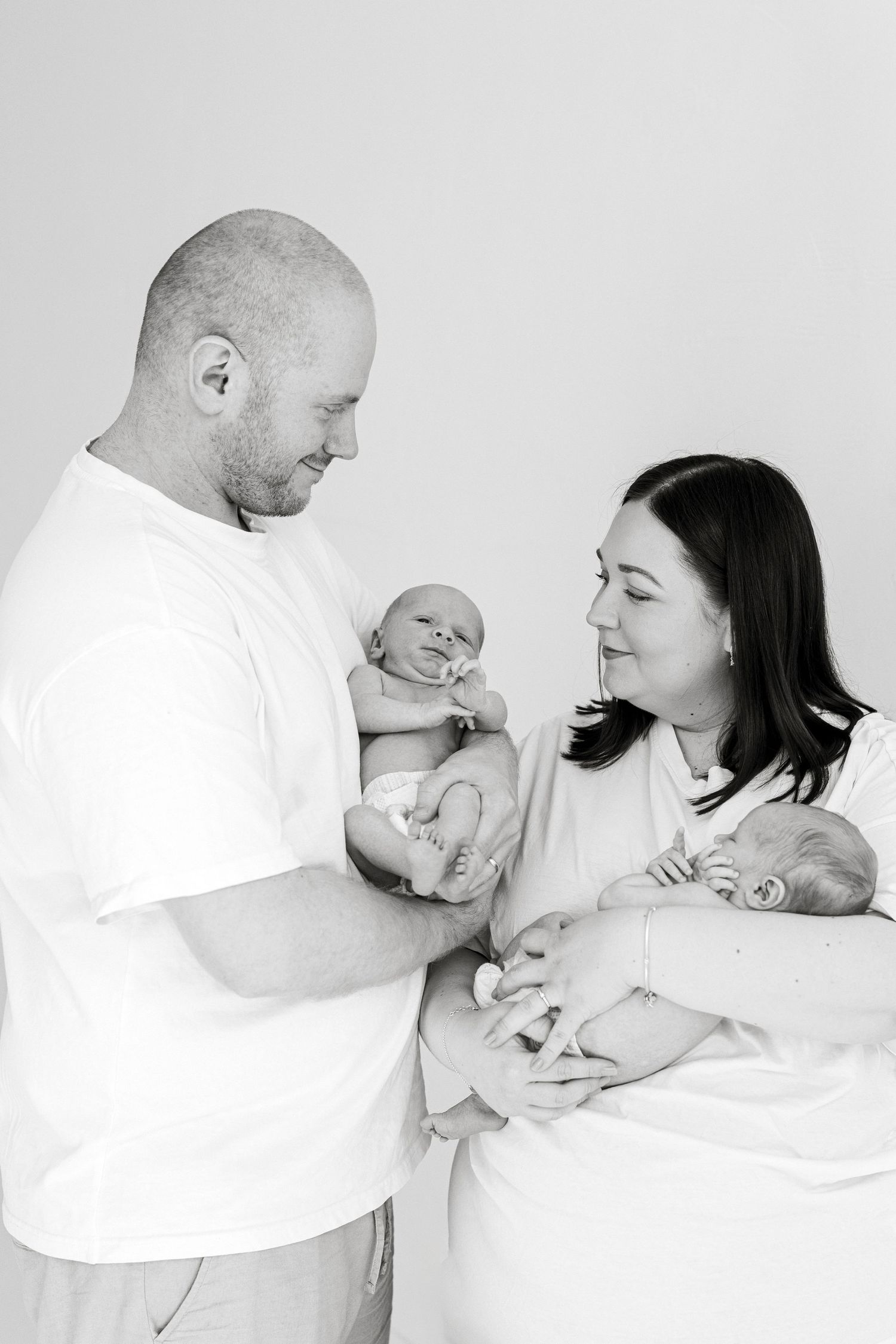 Black and white photo of parents holding newborn twins, smiling lovingly at their babies in a bright studio setting.