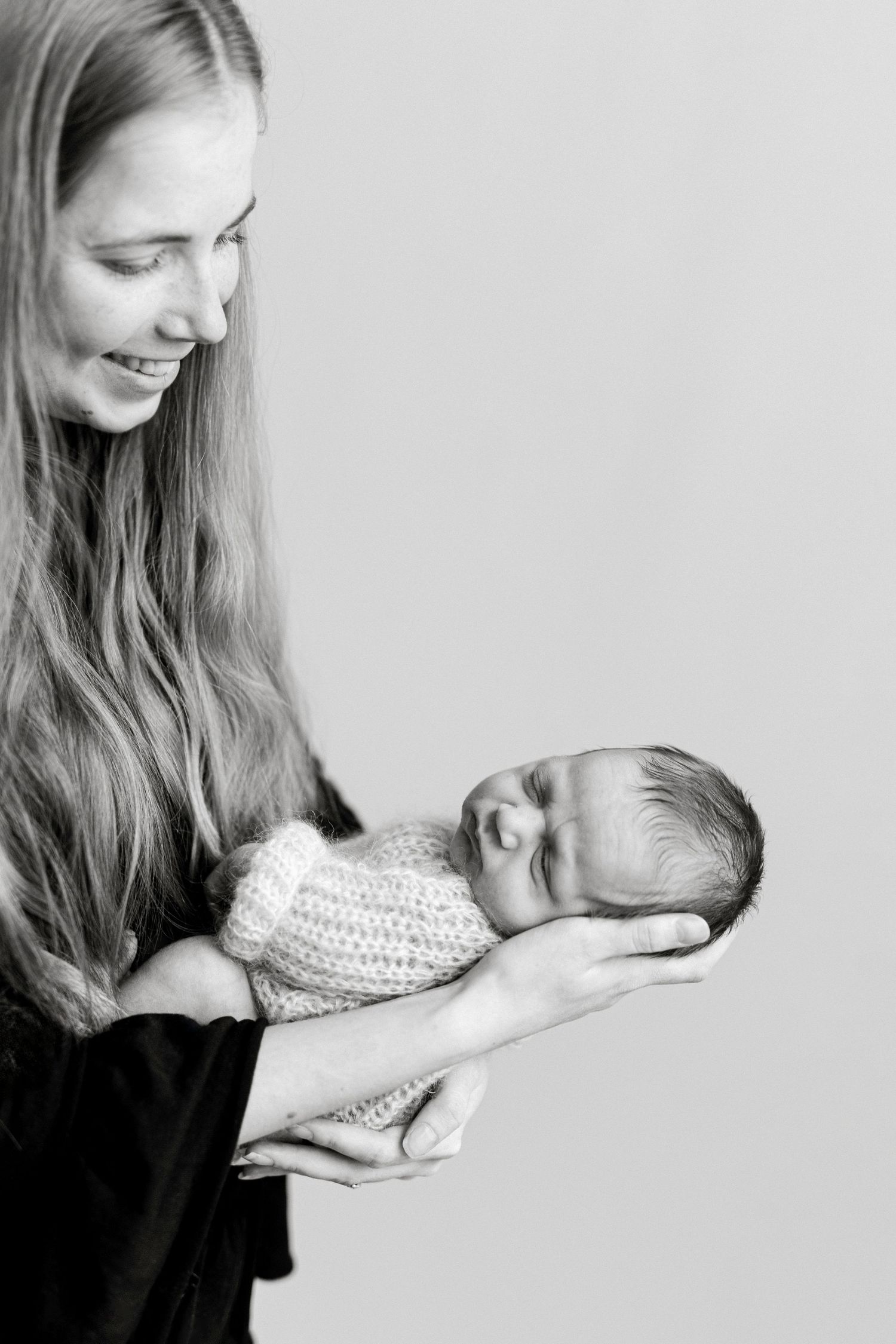 Black and white photo of a smiling mother with long hair cradling her sleeping newborn baby wrapped in a knit blanket.