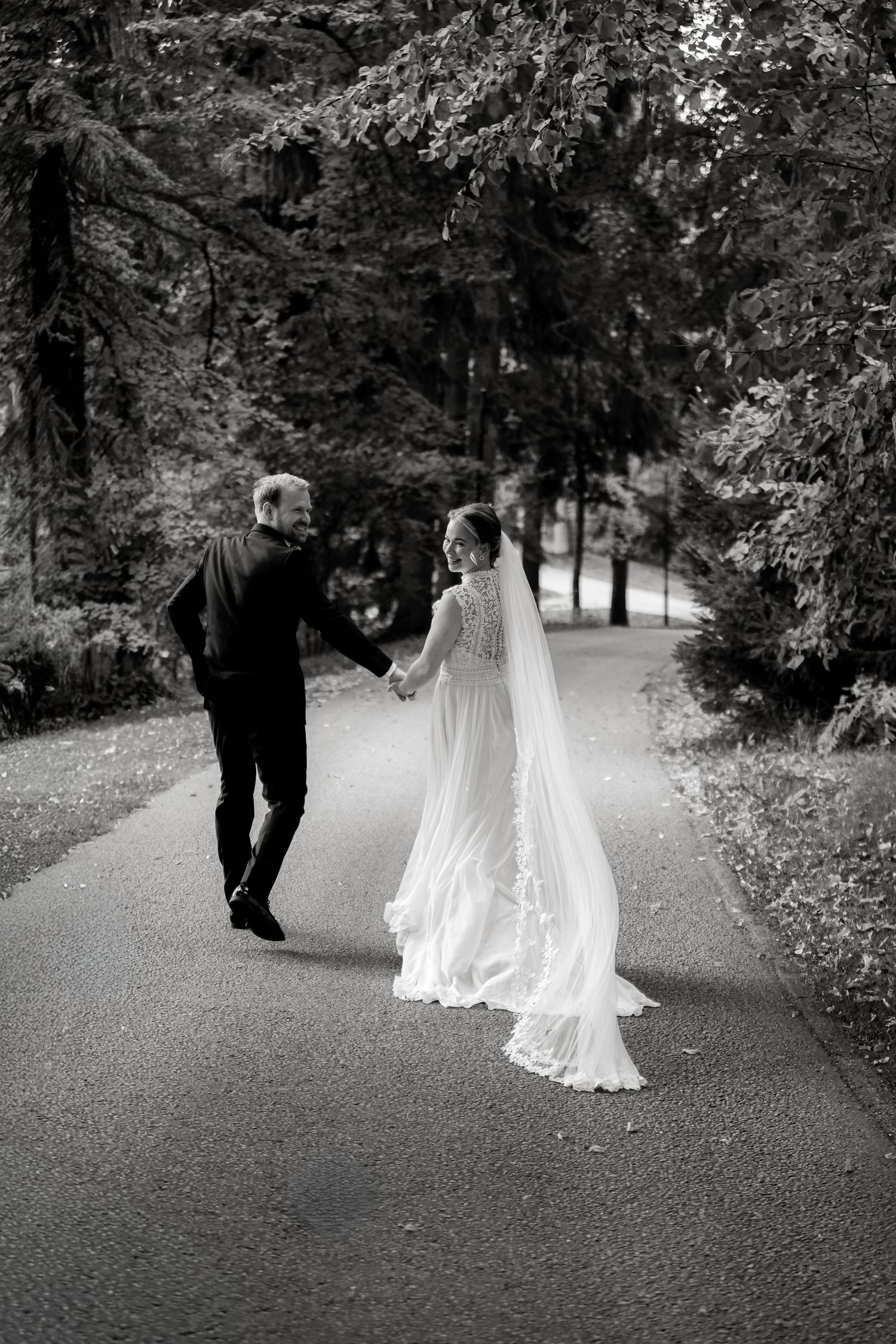 Bride and groom walking hand in hand down tree-lined path in black and white.