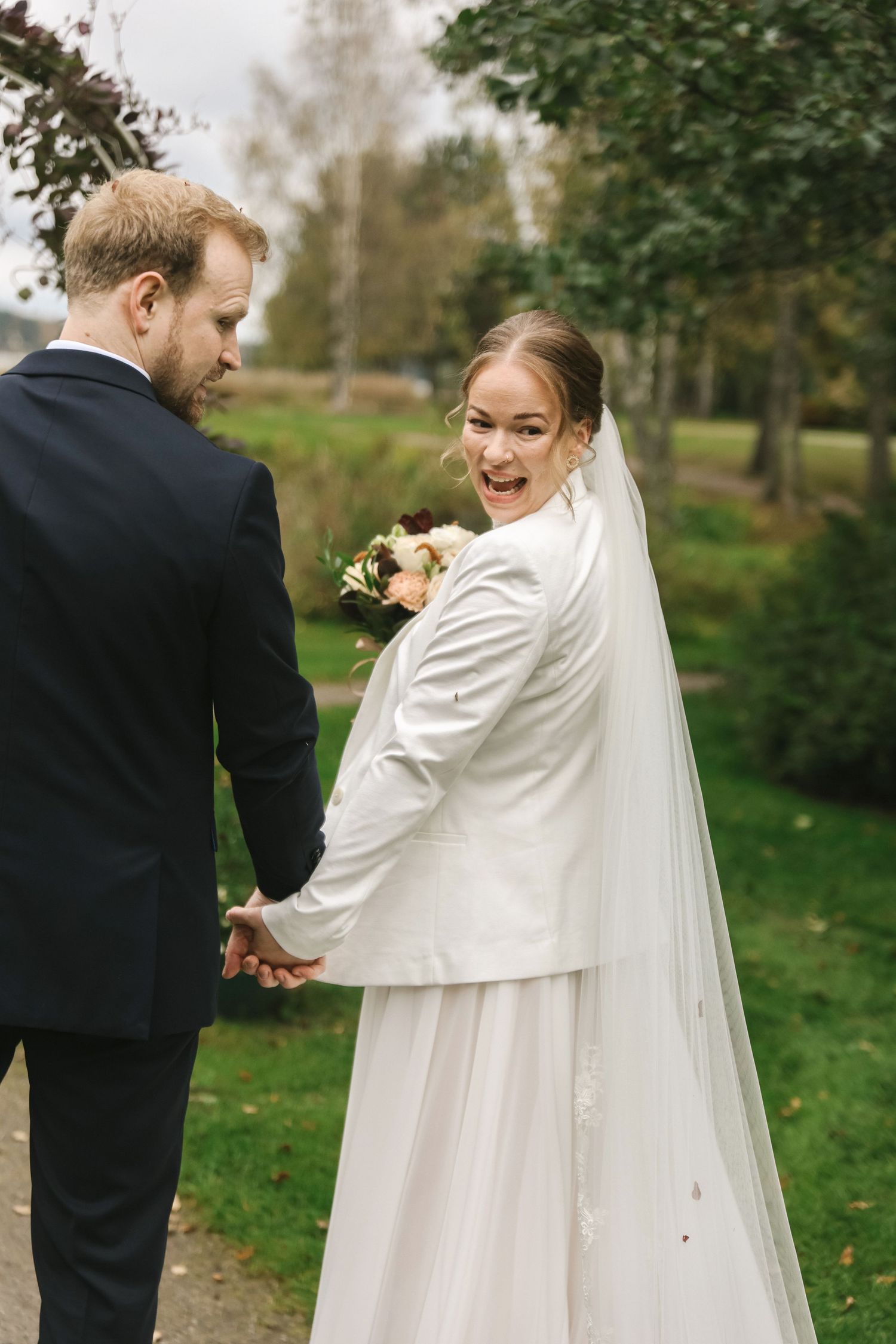 Bride in white dress and veil holds groom's hand, glancing back with a smile in a lush green outdoor setting.
