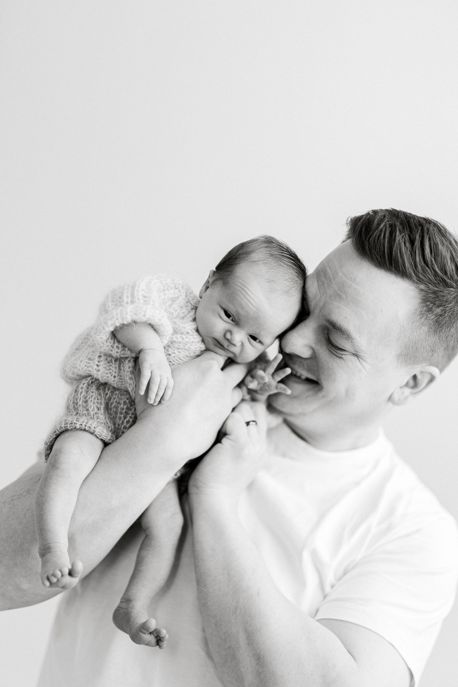 Black and white photo of a smiling man holding a newborn baby in a knit outfit close to his face.