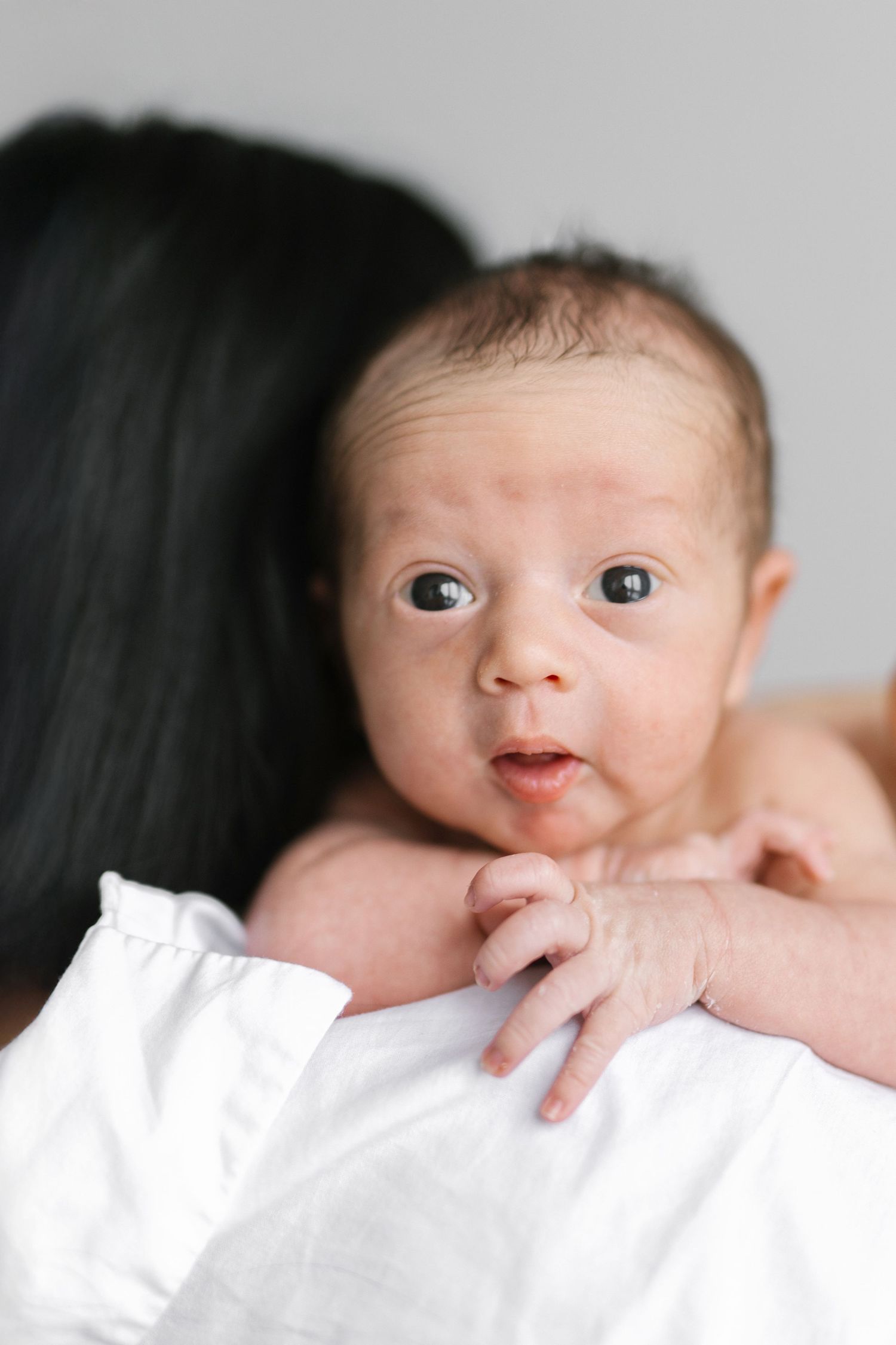 Newborn baby peeking over white blanket with bright curious eyes.