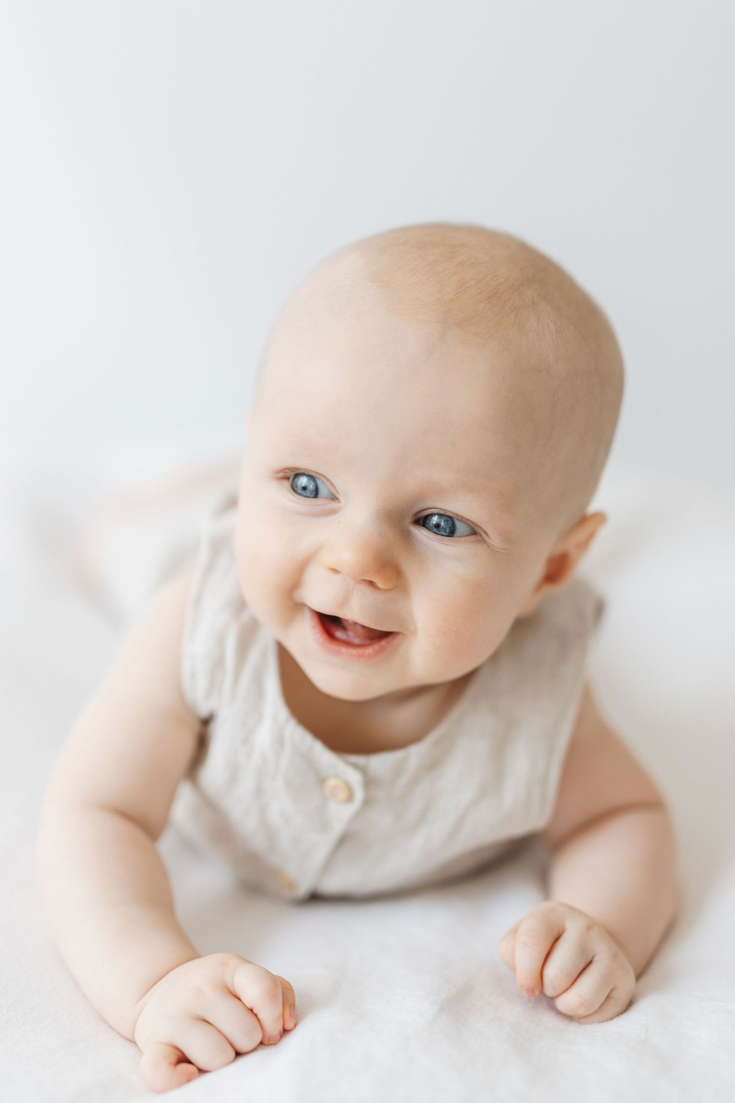 Smiling baby with blue eyes lying on tummy wearing a light gray romper on a white fluffy blanket.