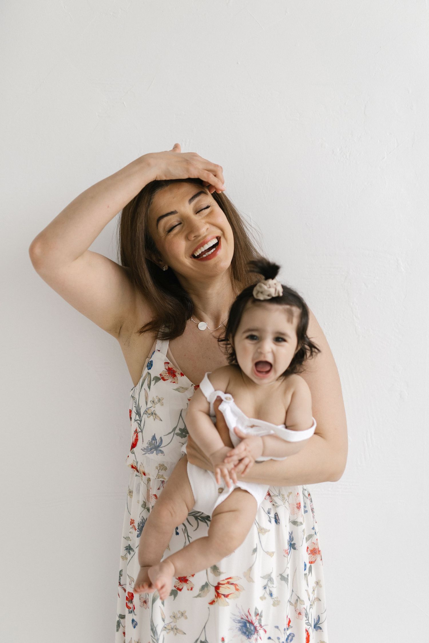 A laughing mother in a floral dress holds her baby girl, both wearing matching outfits against a white background.