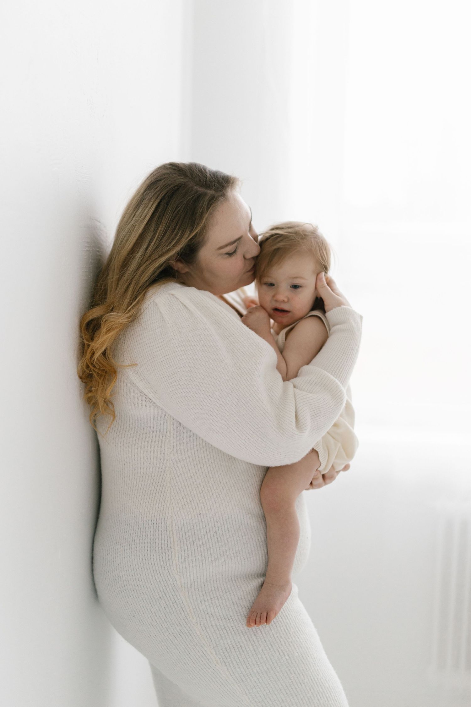 A mother in a white dress tenderly kisses her baby while leaning against a bright white wall.