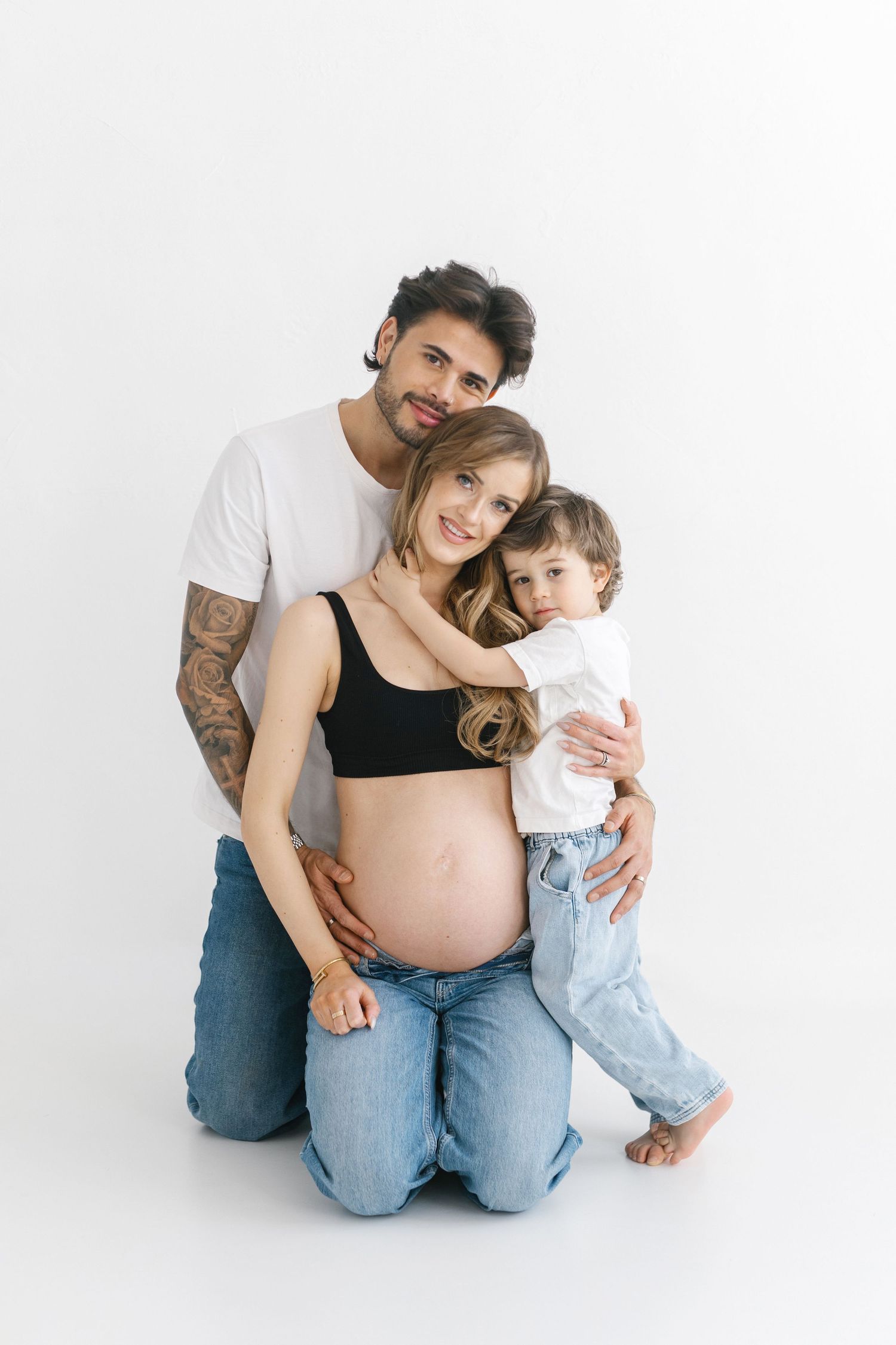 Modern family portrait with pregnant mother, father and young child against white backdrop.