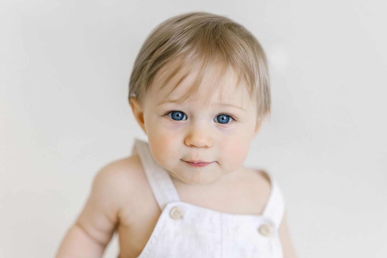 Adorable baby with blue eyes wearing a white linen romper, photographed against a soft white background.