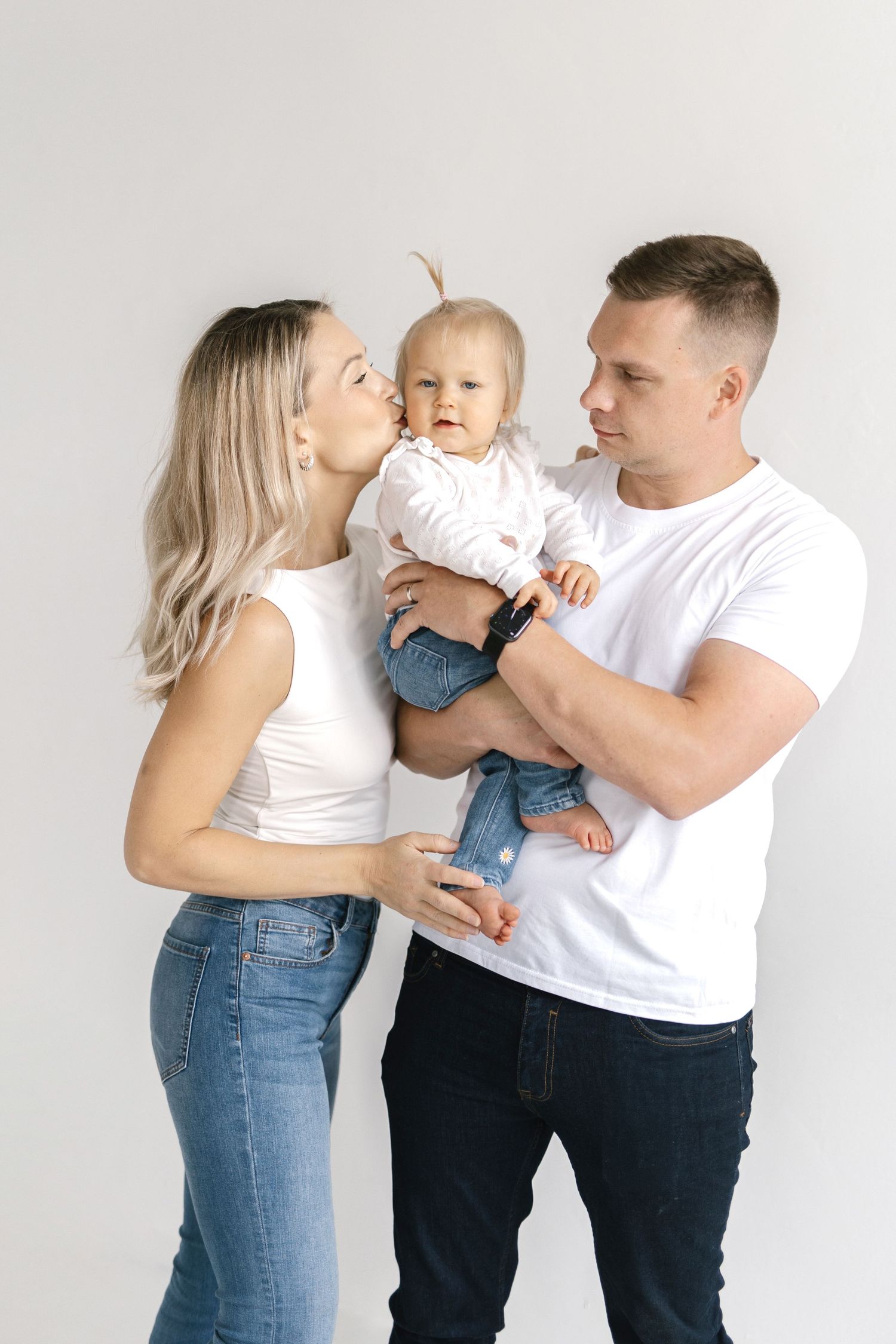A young family in white tops and jeans poses together, with mom kissing baby while dad holds the child.