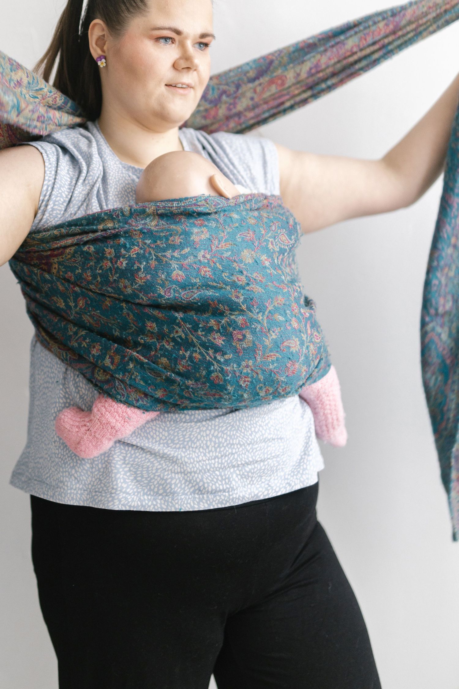 Woman wearing a teal floral baby wrap carrier with infant, adjusting the wrap fabric while standing against white background.