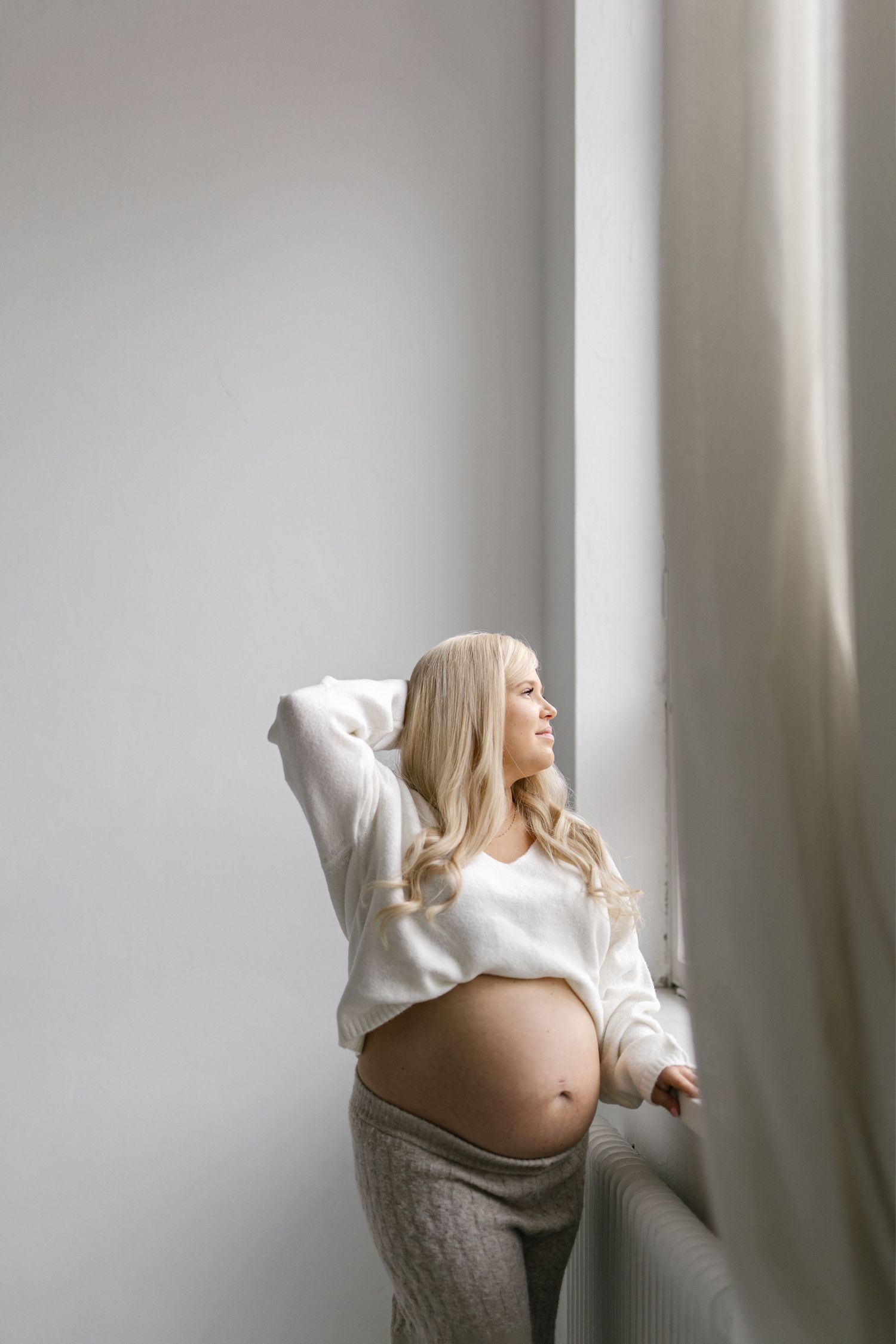 Smiling pregnant woman with blonde hair stands by window in white crop sweater and gray pants, cradling her baby bump.