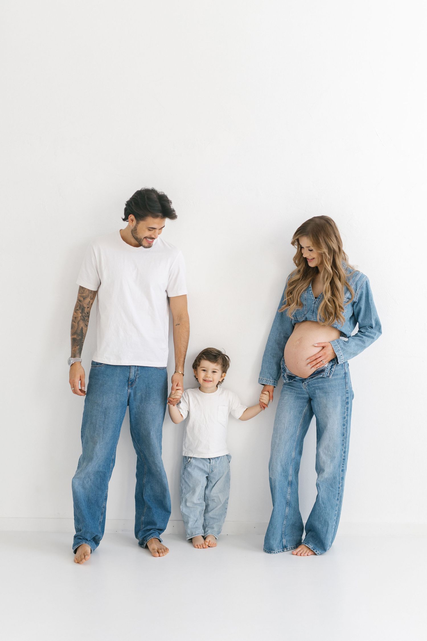Pregnant woman in denim holds hands with toddler and tattooed partner against white background.