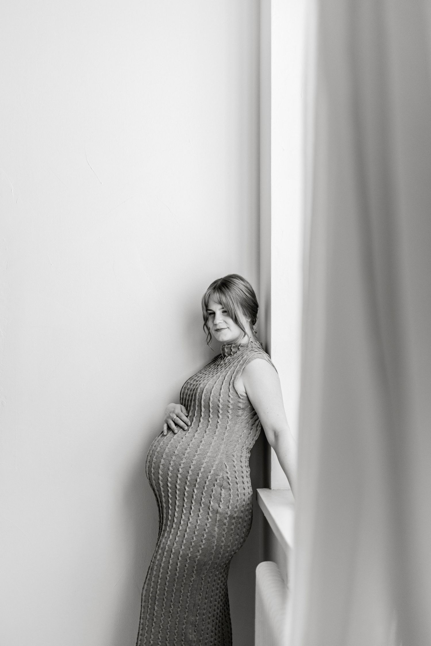 Black and white photo of a pregnant woman in a knit dress leaning against a wall, cradling her baby bump.
