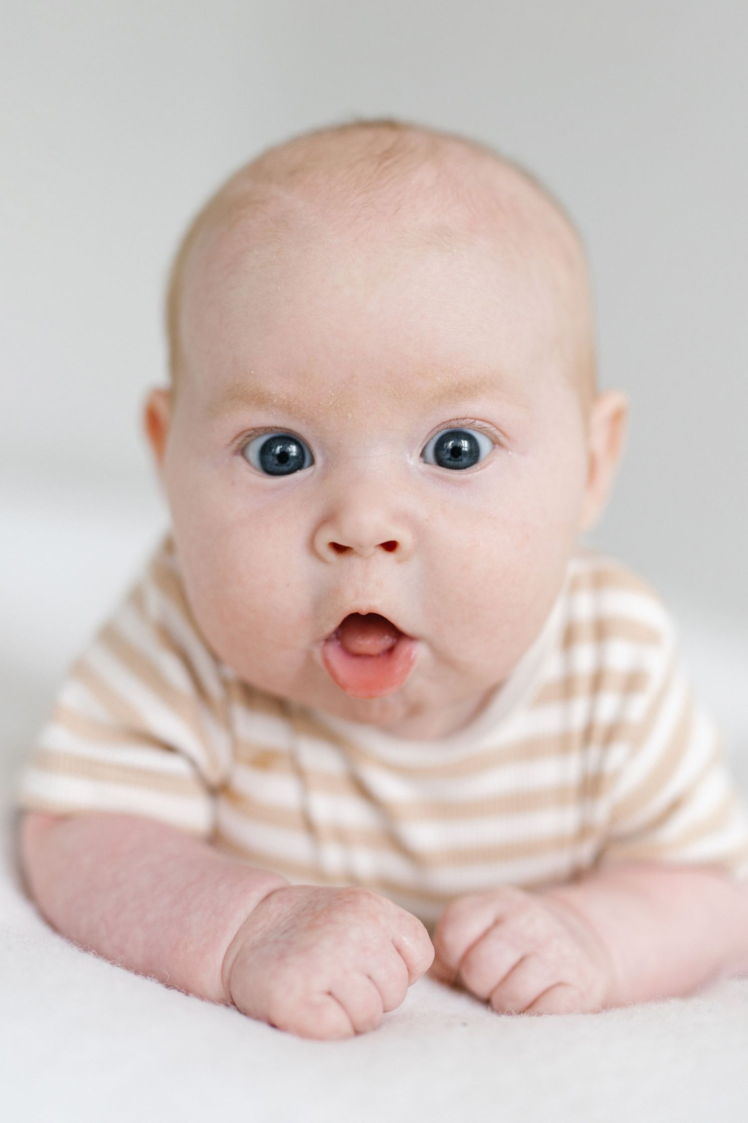 Adorable baby with blue eyes lying on tummy wearing striped onesie, looking surprised with mouth open.