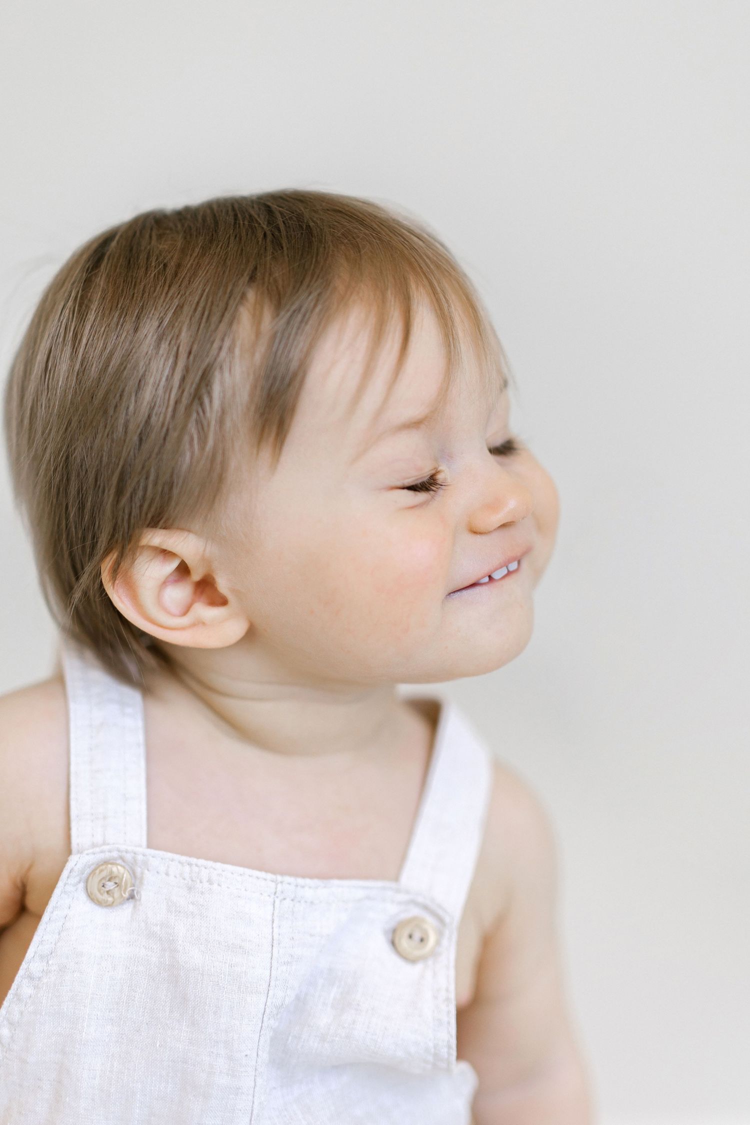 Smiling baby toddler in white linen overalls against a light background, looking to the side with a cheerful expression.