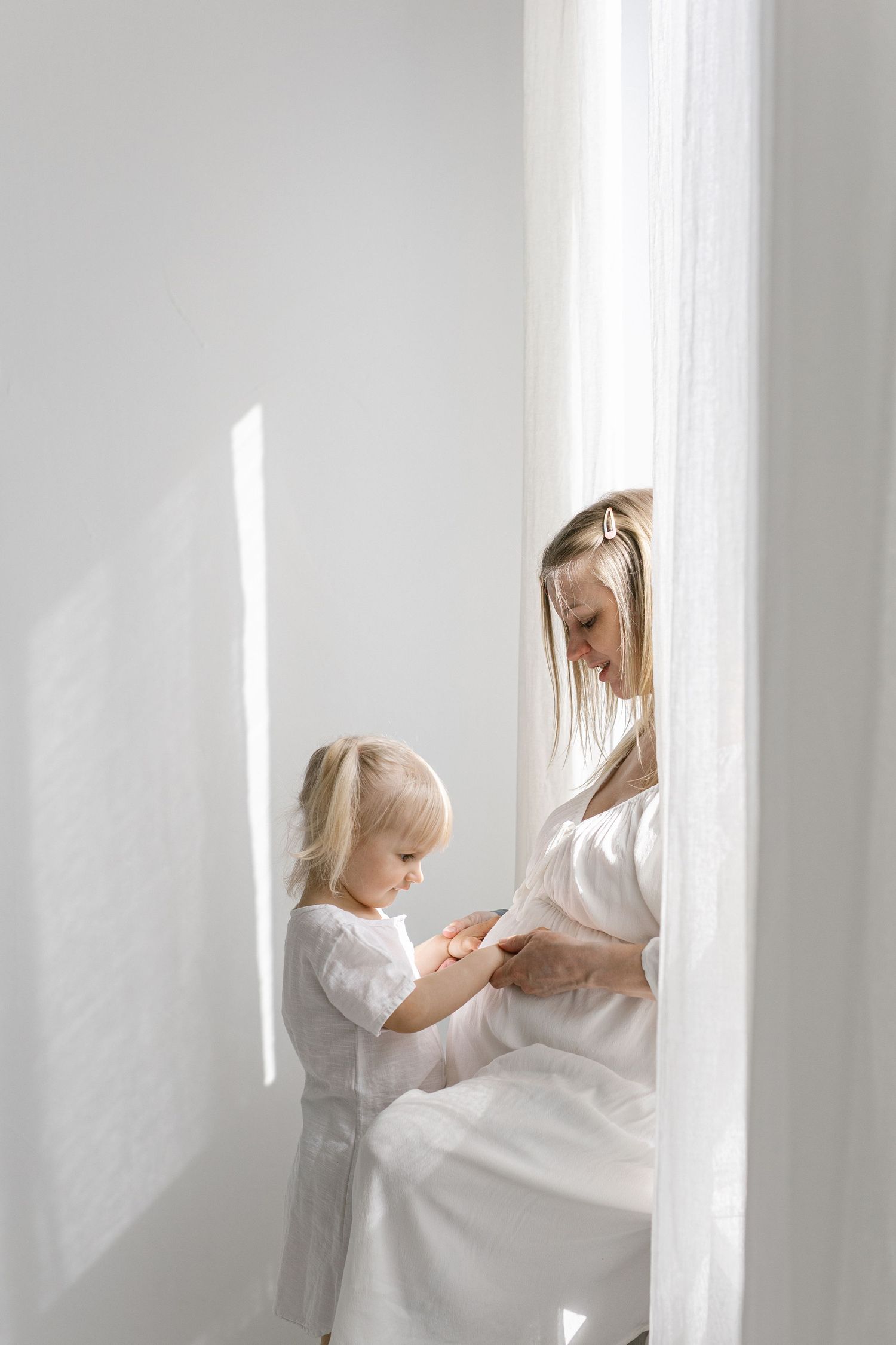 A pregnant woman in white dress tenderly interacts with her toddler daughter near a sunlit white curtain.