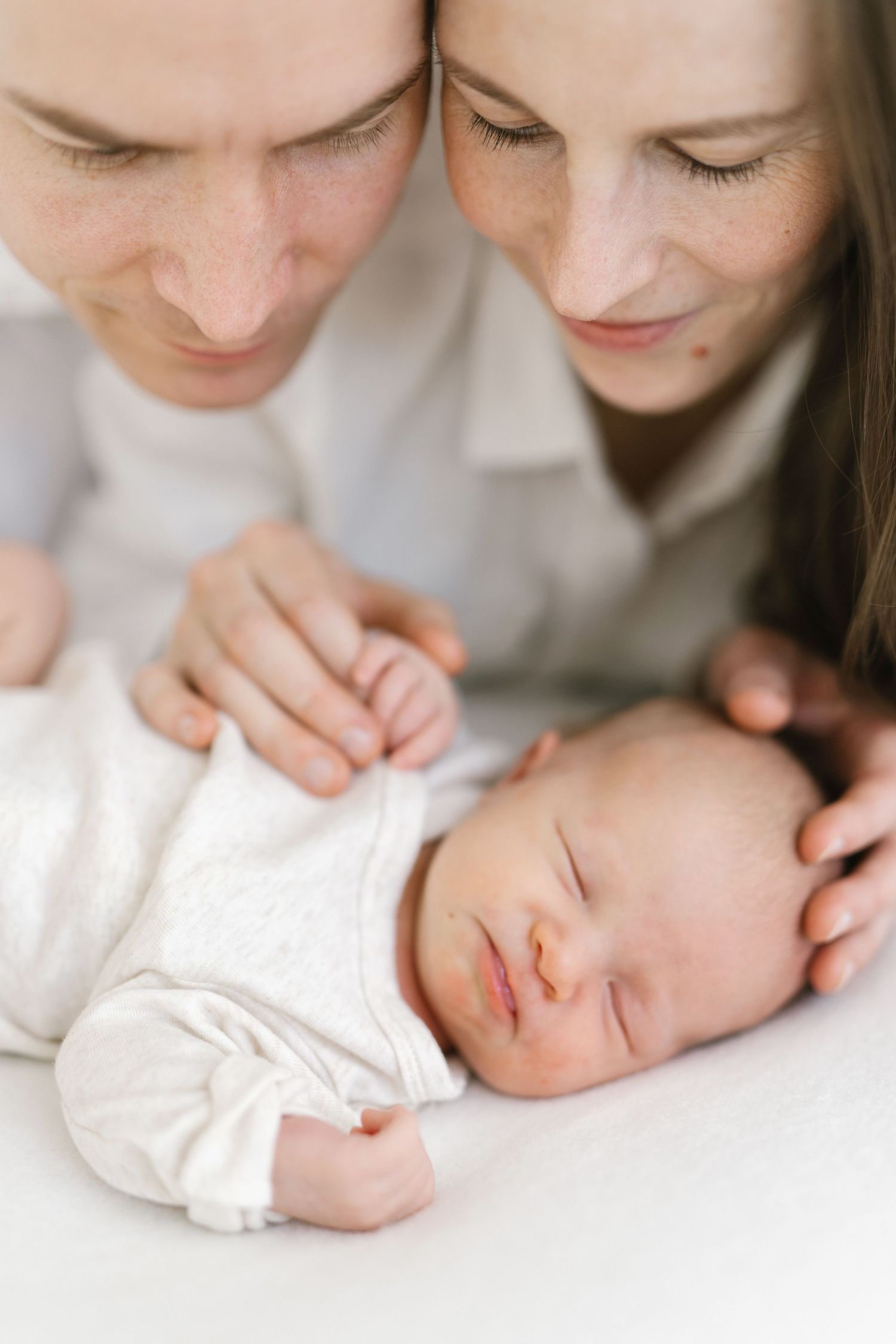 Two parents lean over their sleeping newborn baby dressed in white, sharing a tender moment together.