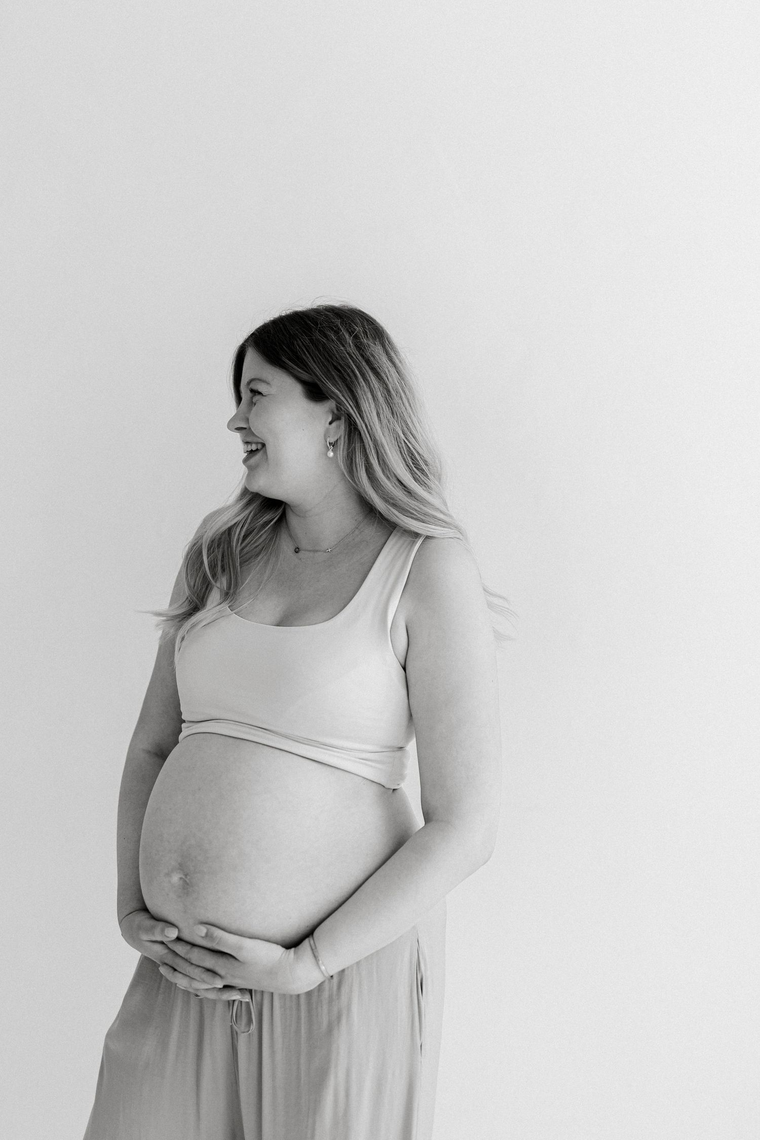 Black and white maternity portrait of a smiling pregnant woman in a crop top and loose pants, cradling her bare belly against a plain studio