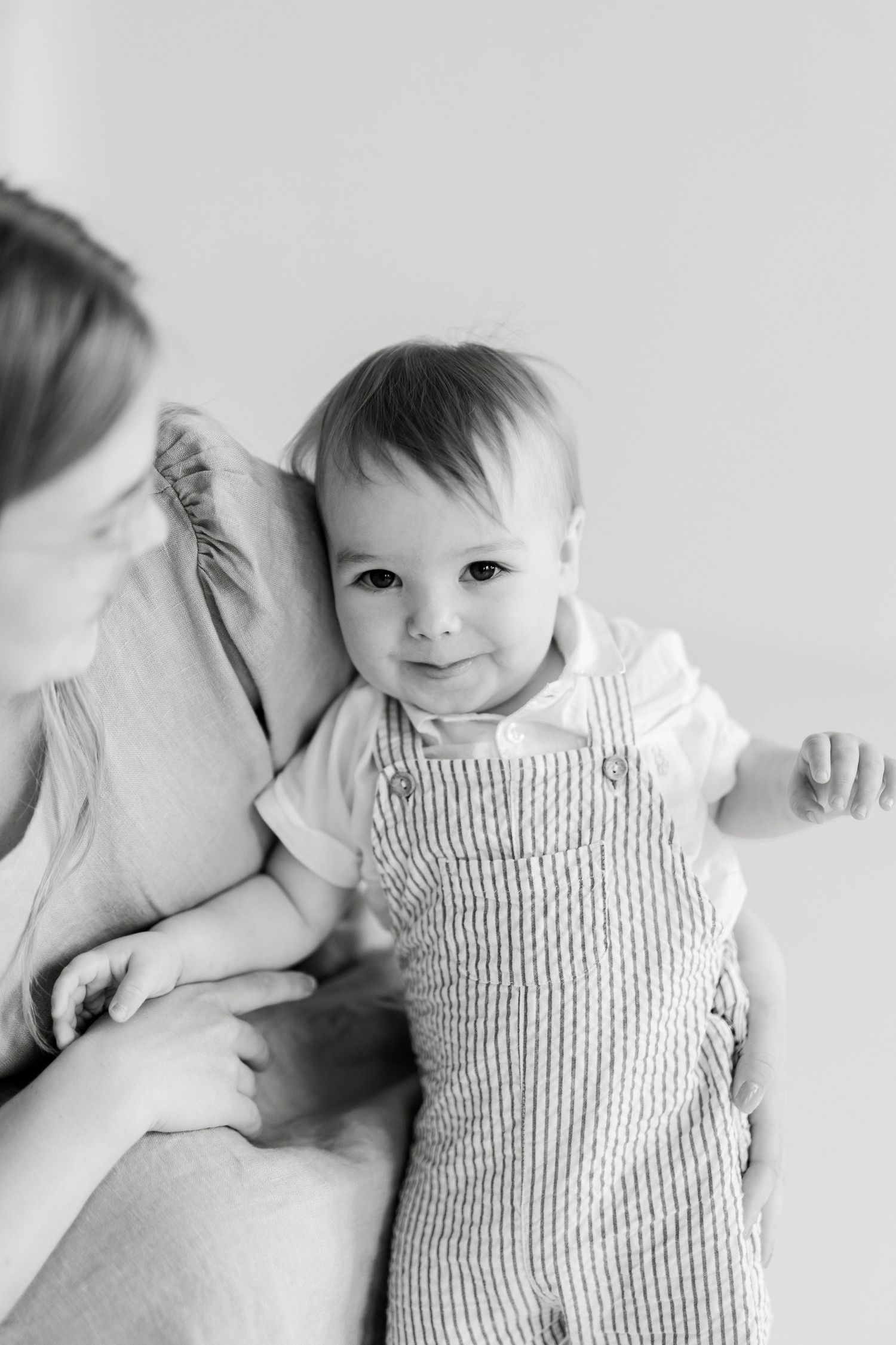 Black and white photo of a smiling baby in striped overalls being held by a mother in a bright studio setting.