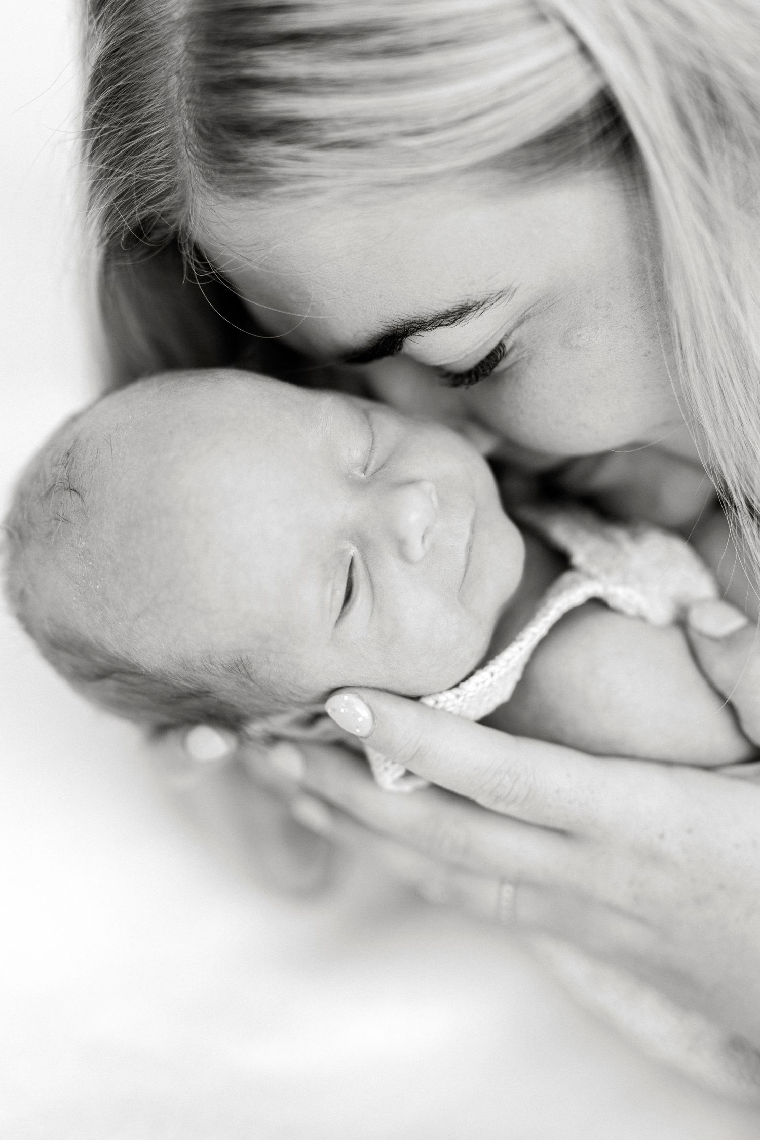 Black and white photo of a mother tenderly kissing her sleeping newborn baby cradled in her hands.