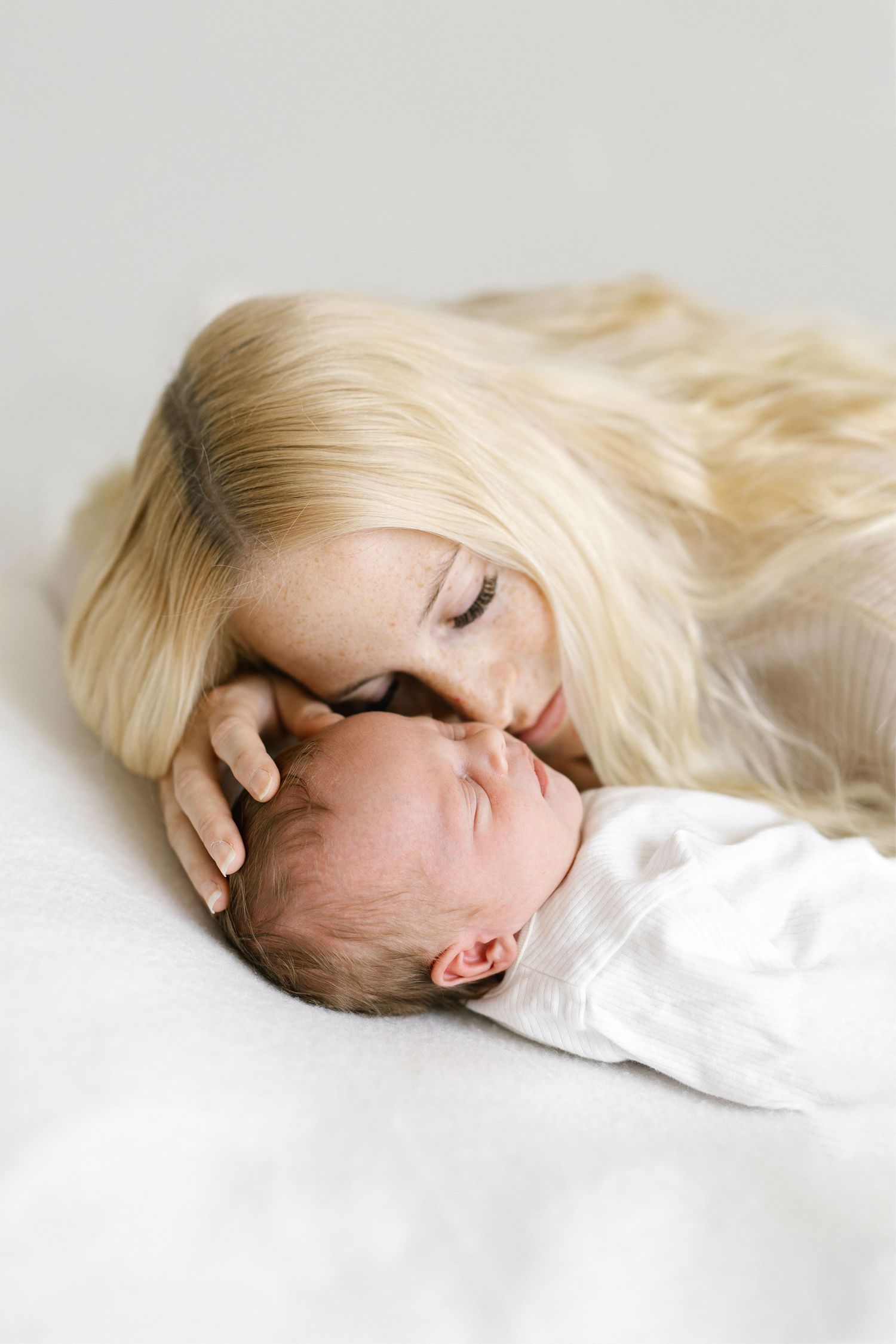 Blonde mother tenderly kissing her sleeping newborn baby wrapped in white, lying together on a soft white surface.