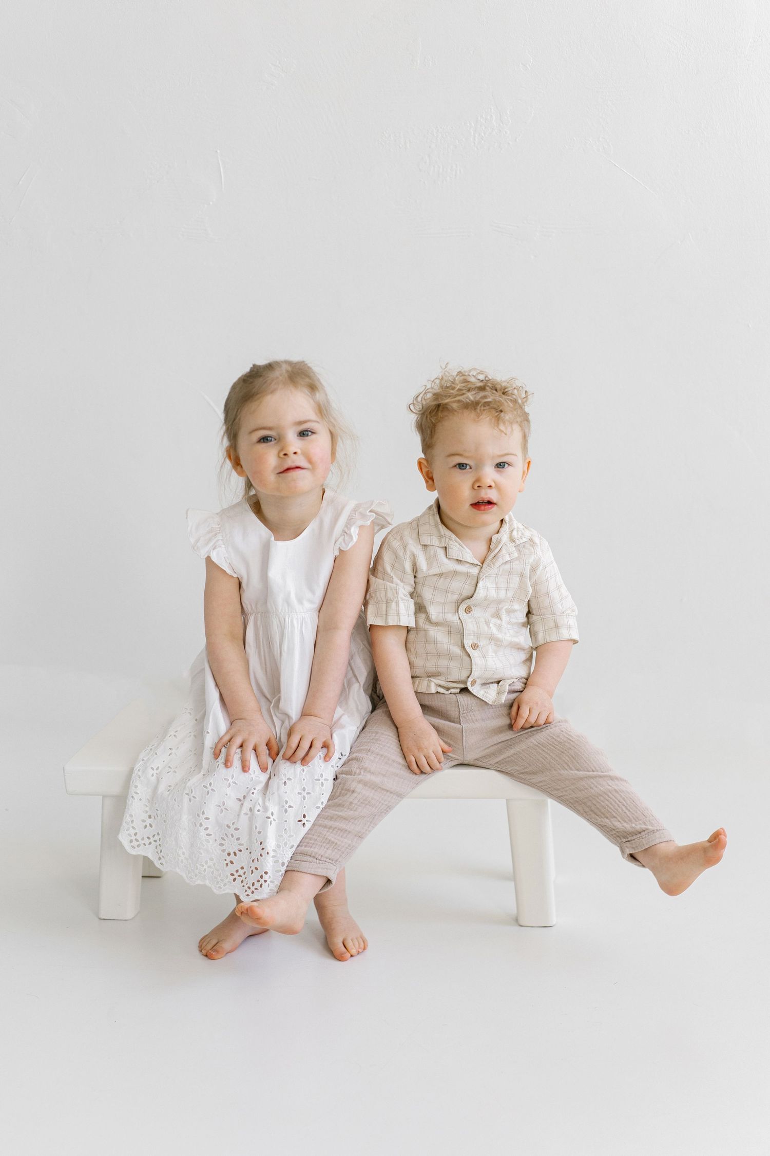 Two young barefoot children sit on a white bench against a light background, dressed in neutral-toned outfits.