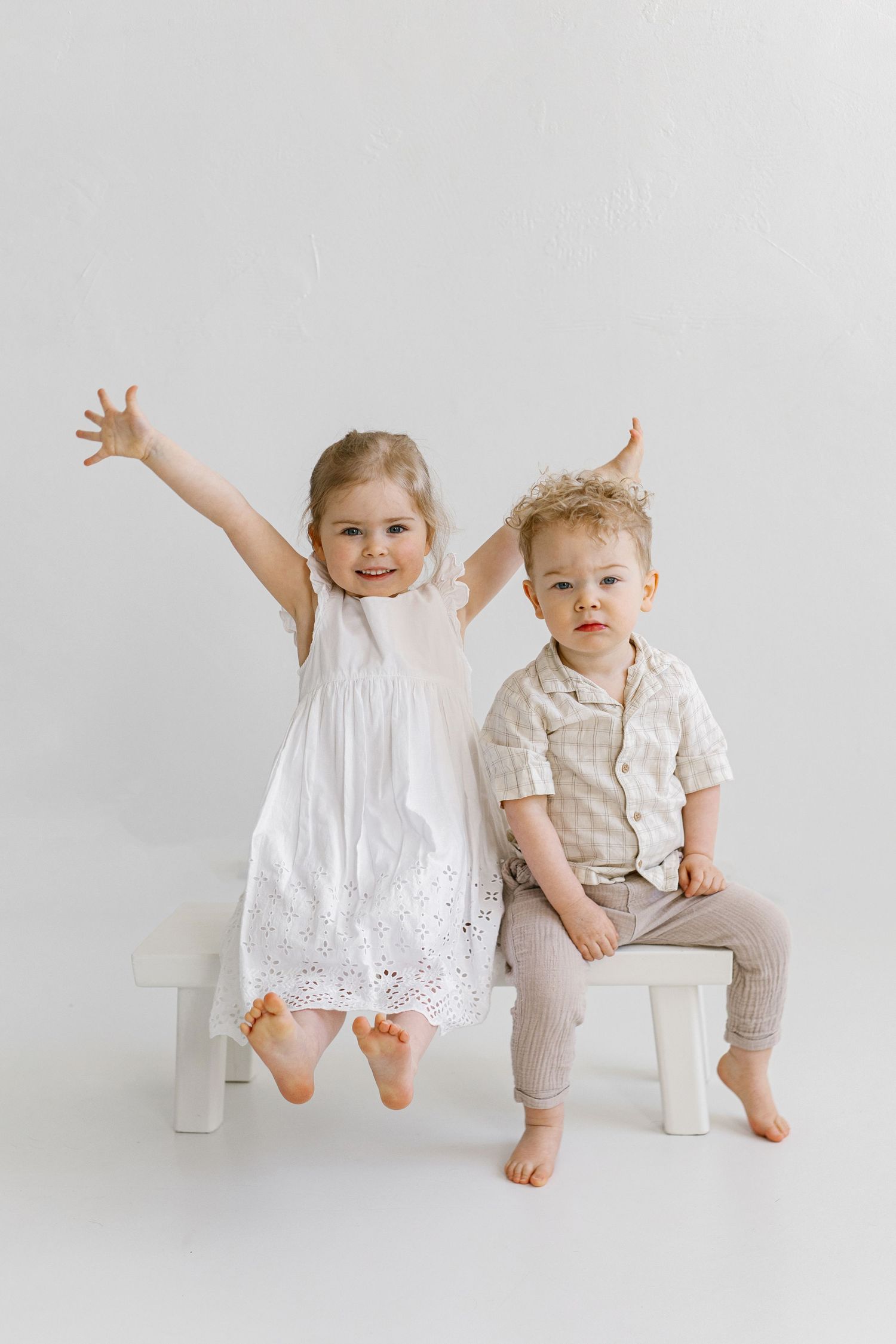 Two young children sitting on white chairs, girl in white dress with arms raised and boy in beige outfit, studio portrait.