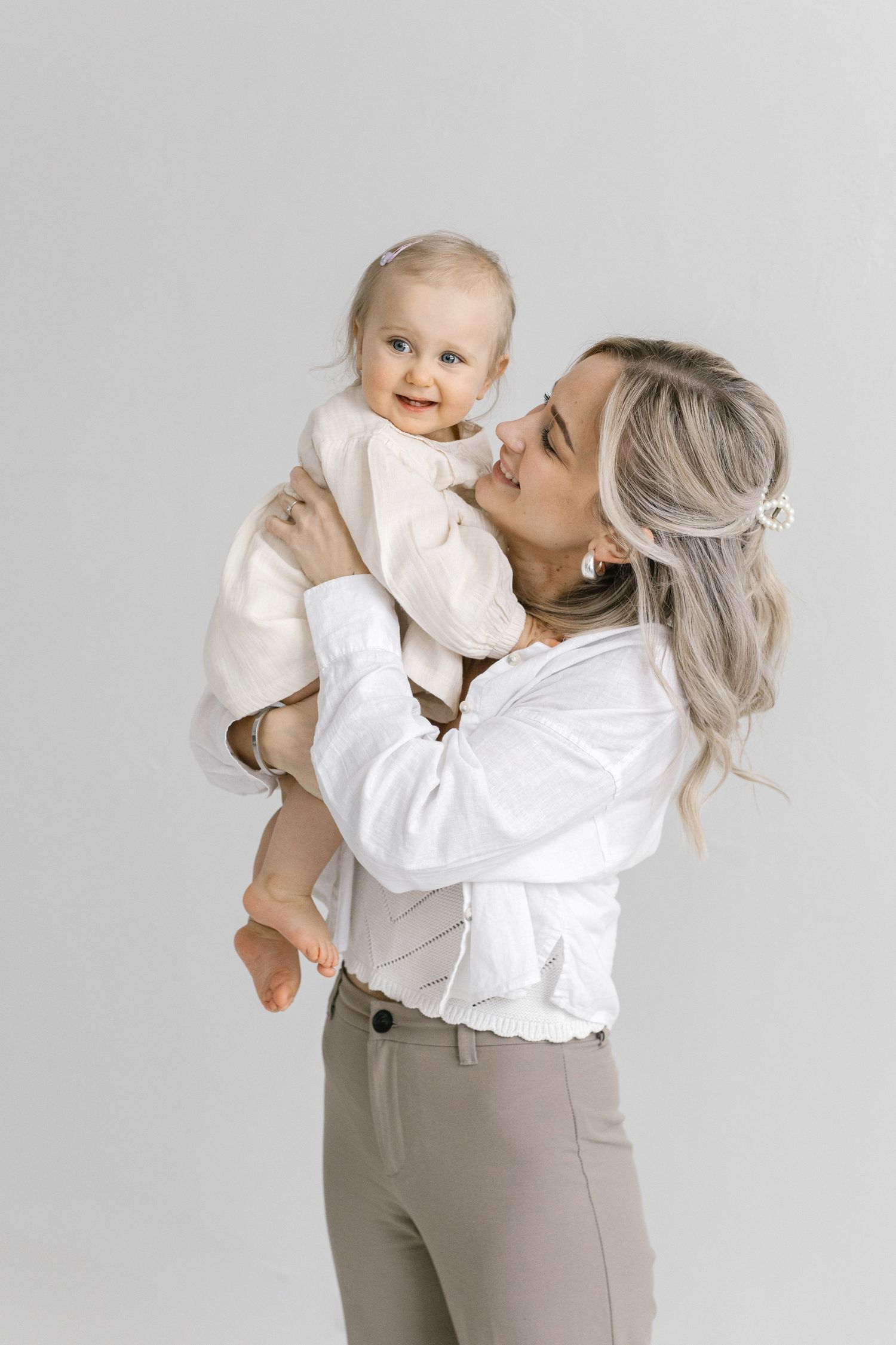 A parent in white top and beige pants lifts and plays with a baby wearing white against a light gray background.