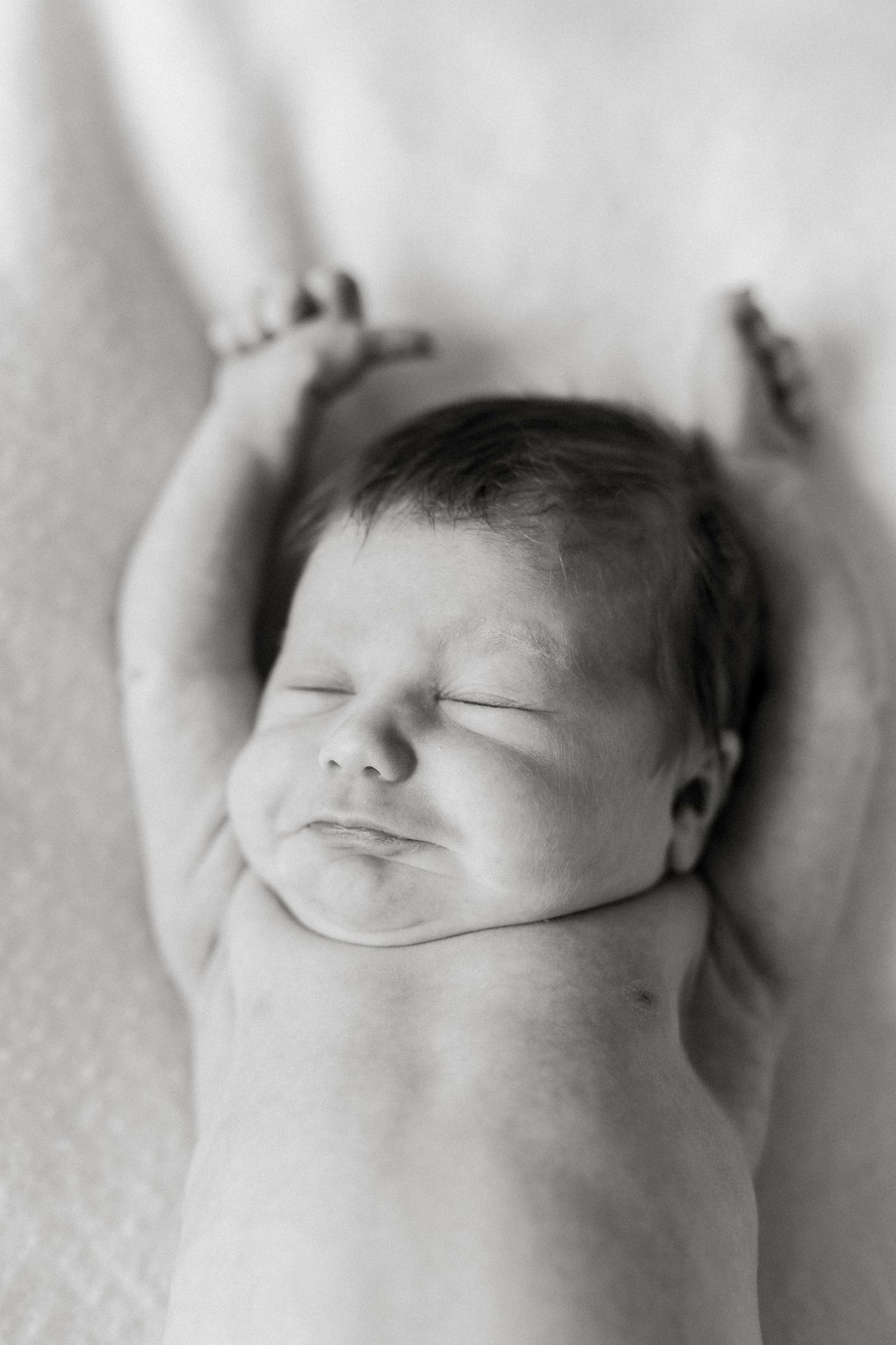 Black and white photo of a sleeping newborn baby held gently by adult hands, arms raised above head.