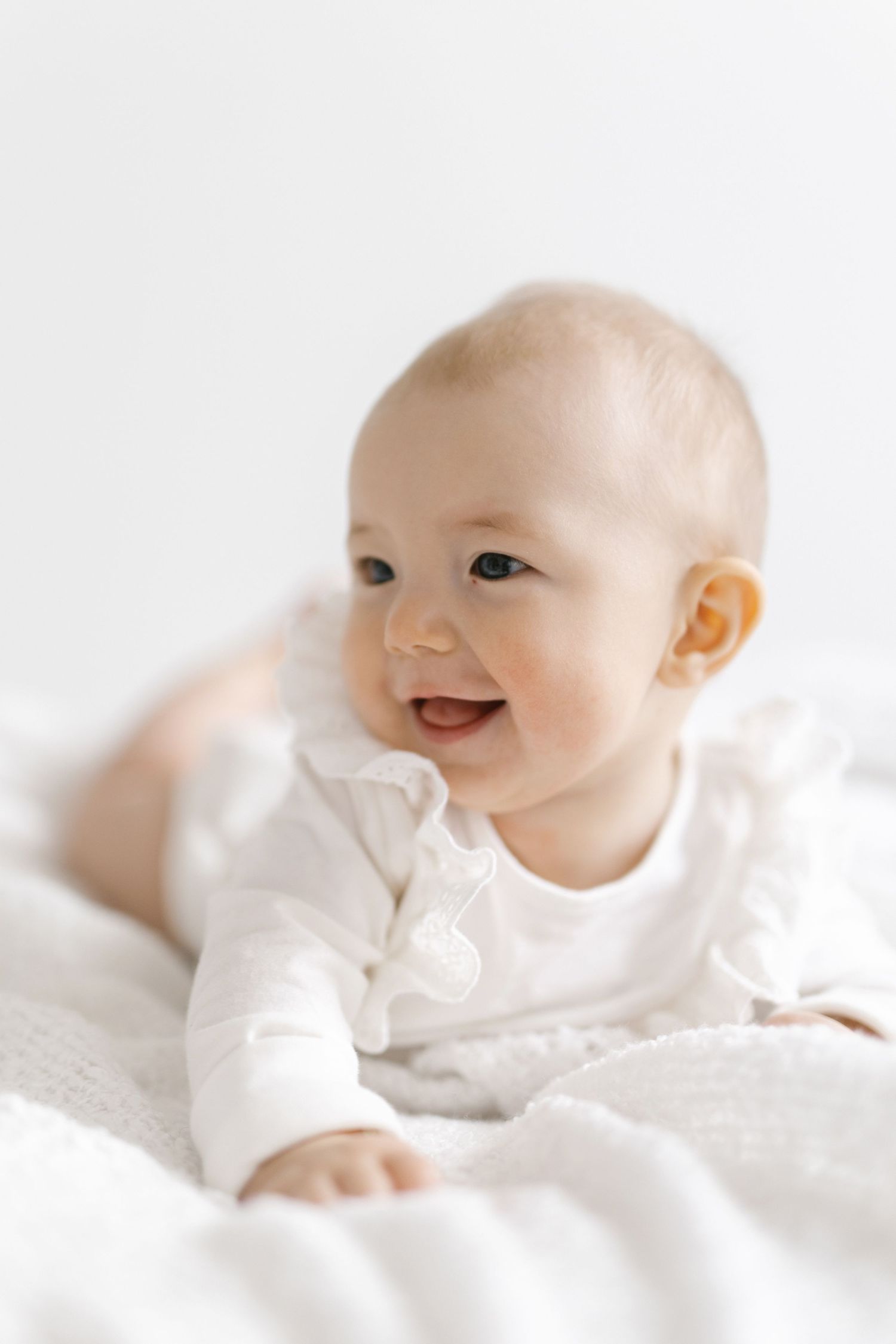 Smiling baby in white outfit doing tummy time on a soft white blanket against a light background.