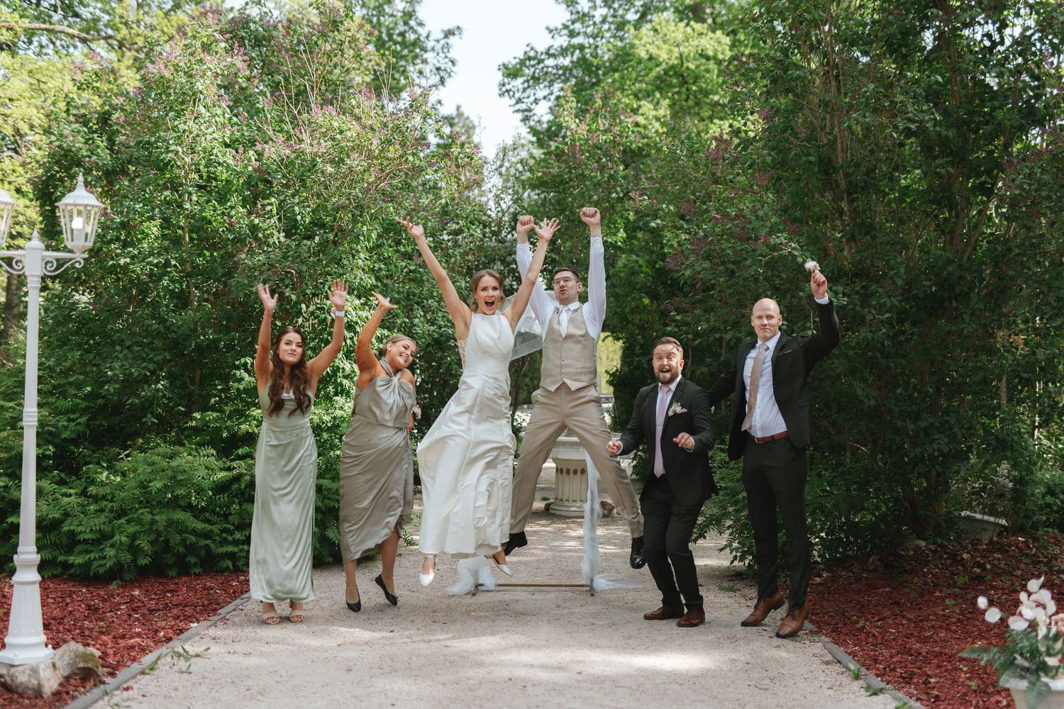 Wedding party jumping joyfully on a garden path surrounded by lush green trees on a bright sunny day.