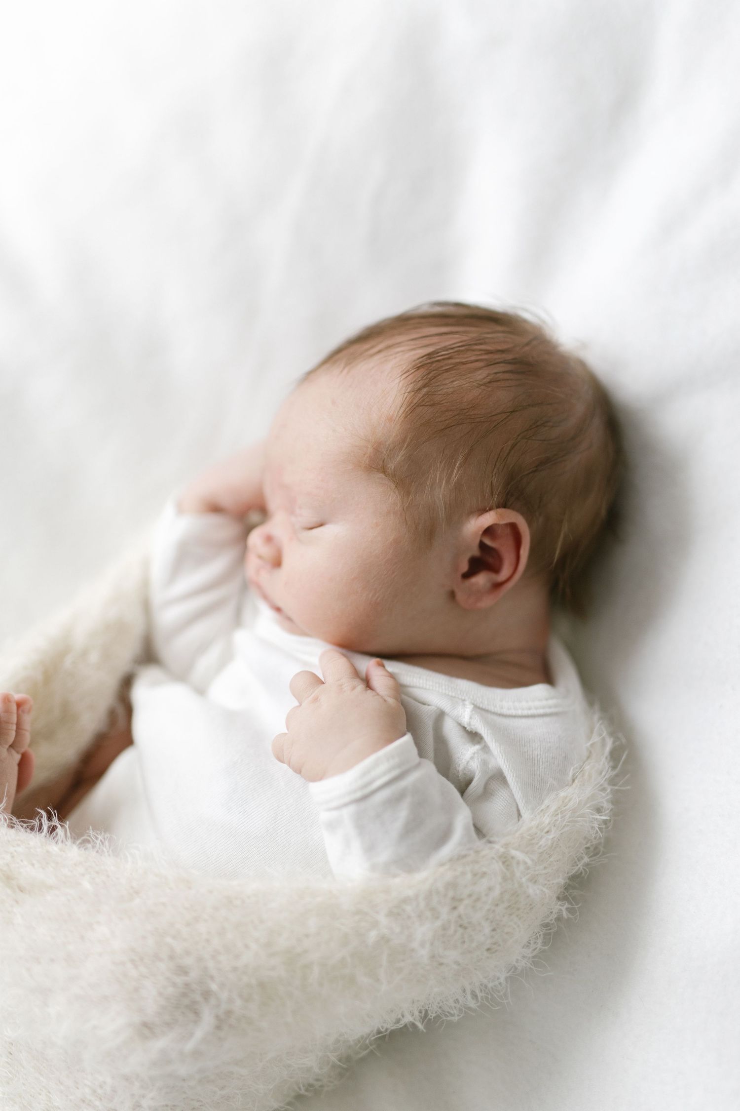 Sleeping newborn in white outfit nestled in fluffy white blanket.