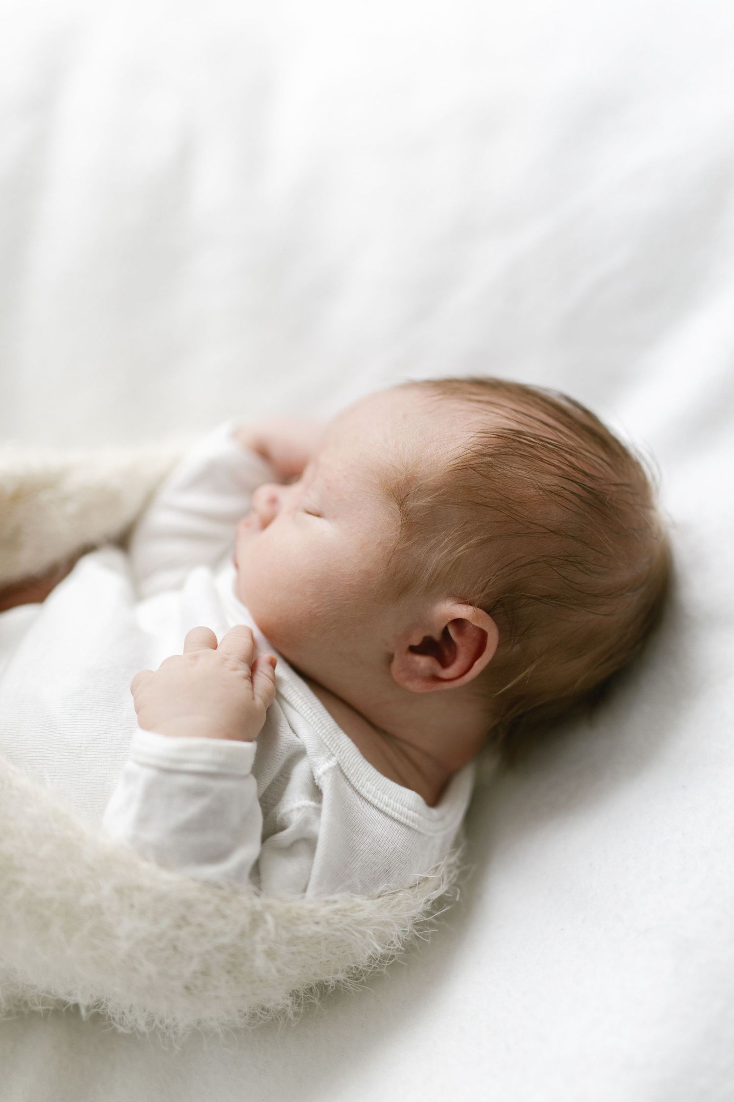 Newborn in white outfit resting on soft white surface.