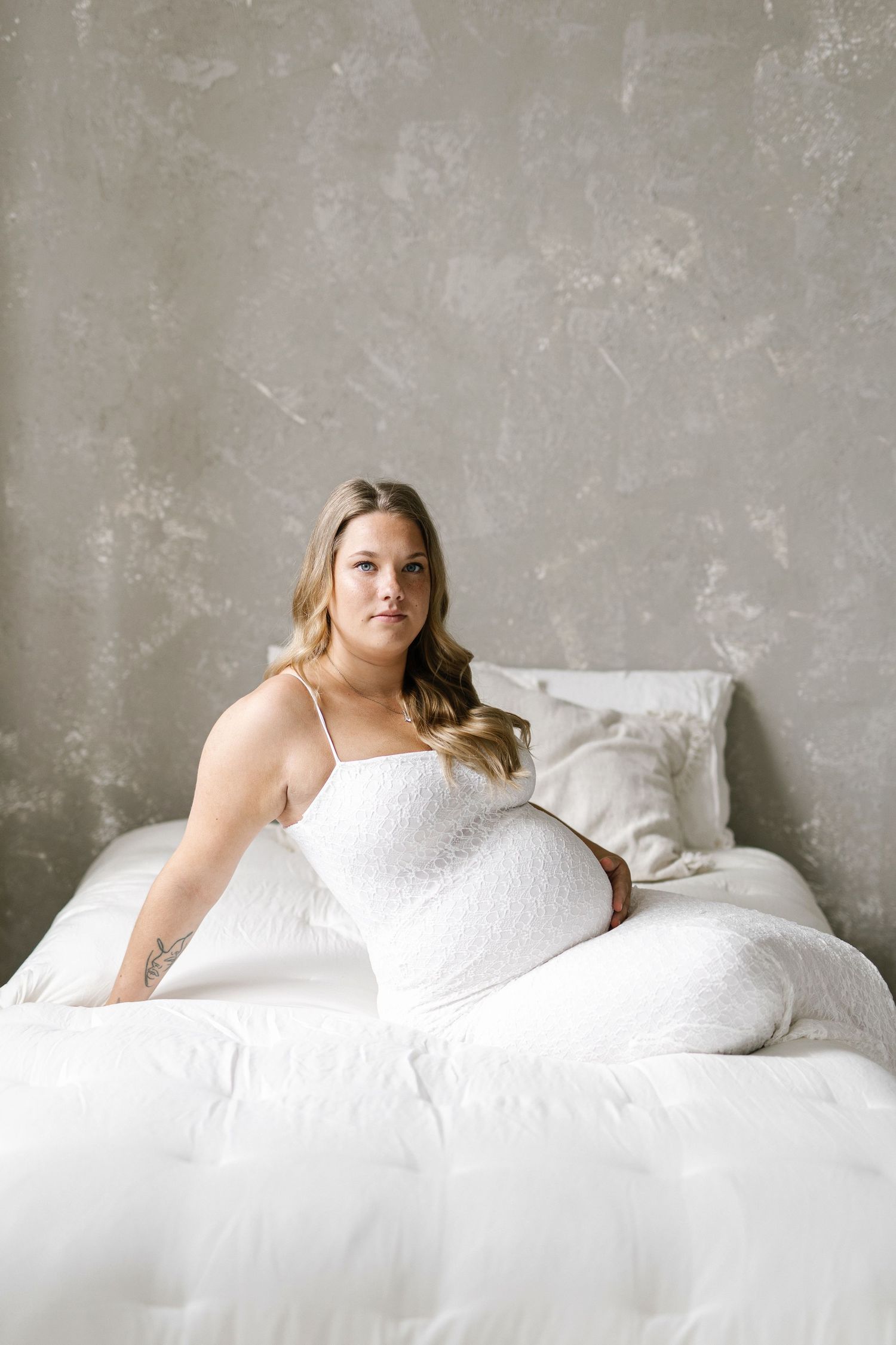 Pregnant woman in white dress sitting on white bed against gray textured wall, maternity photography.