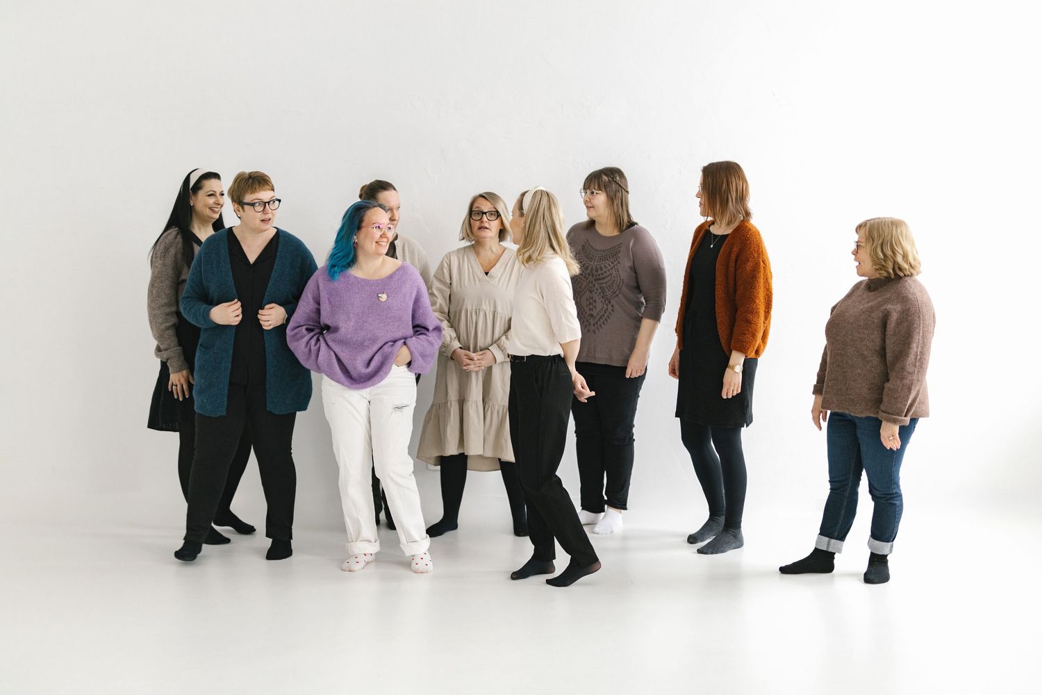Group of eight women posing together in a bright white studio, wearing colorful casual outfits.