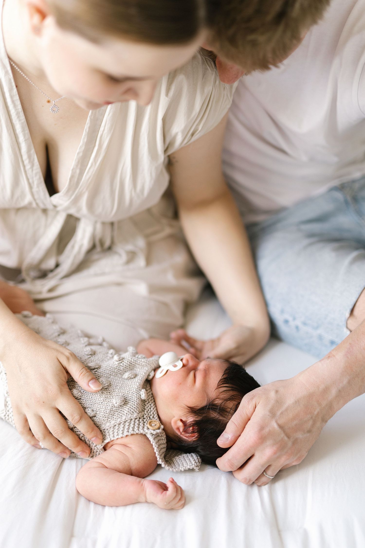 Parents lovingly gaze at their newborn baby lying on a white bed, dressed in a knit outfit with a bow headband.