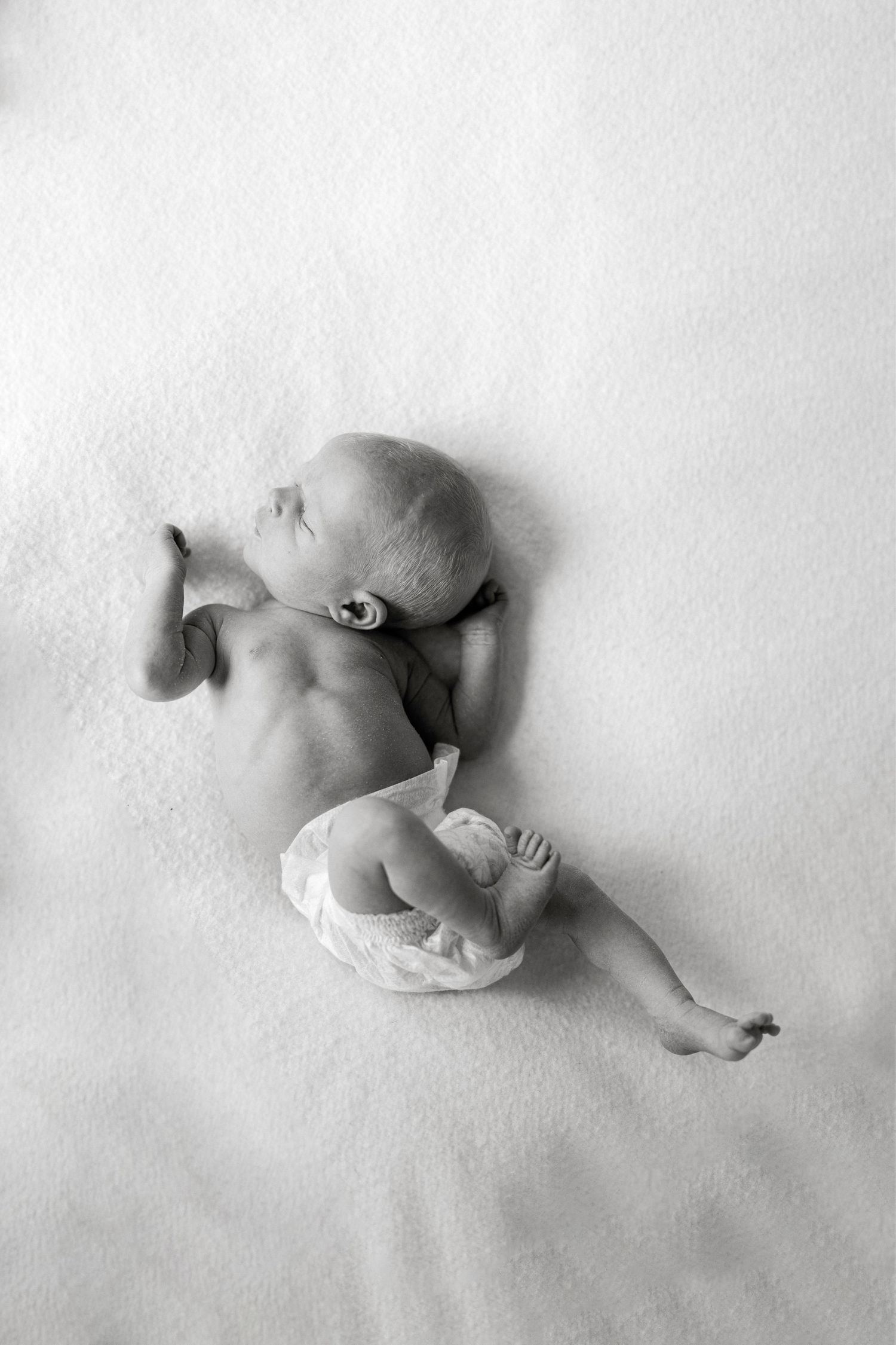Black and white photo of a newborn baby in a diaper lying on a soft white blanket with one arm raised and legs stretched out.