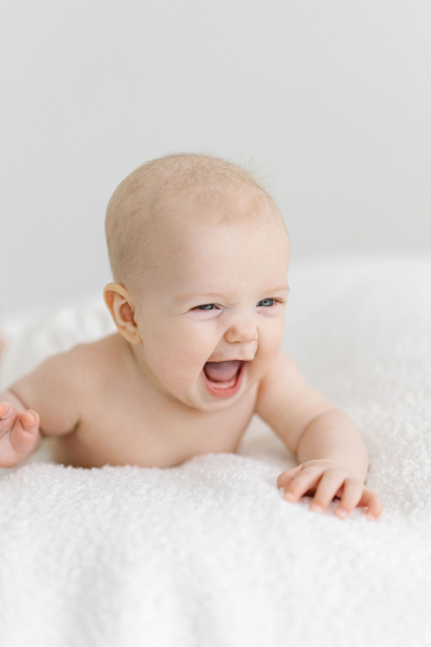 Happy baby lying on white fluffy blanket, laughing with mouth open, tummy time milestone photo.