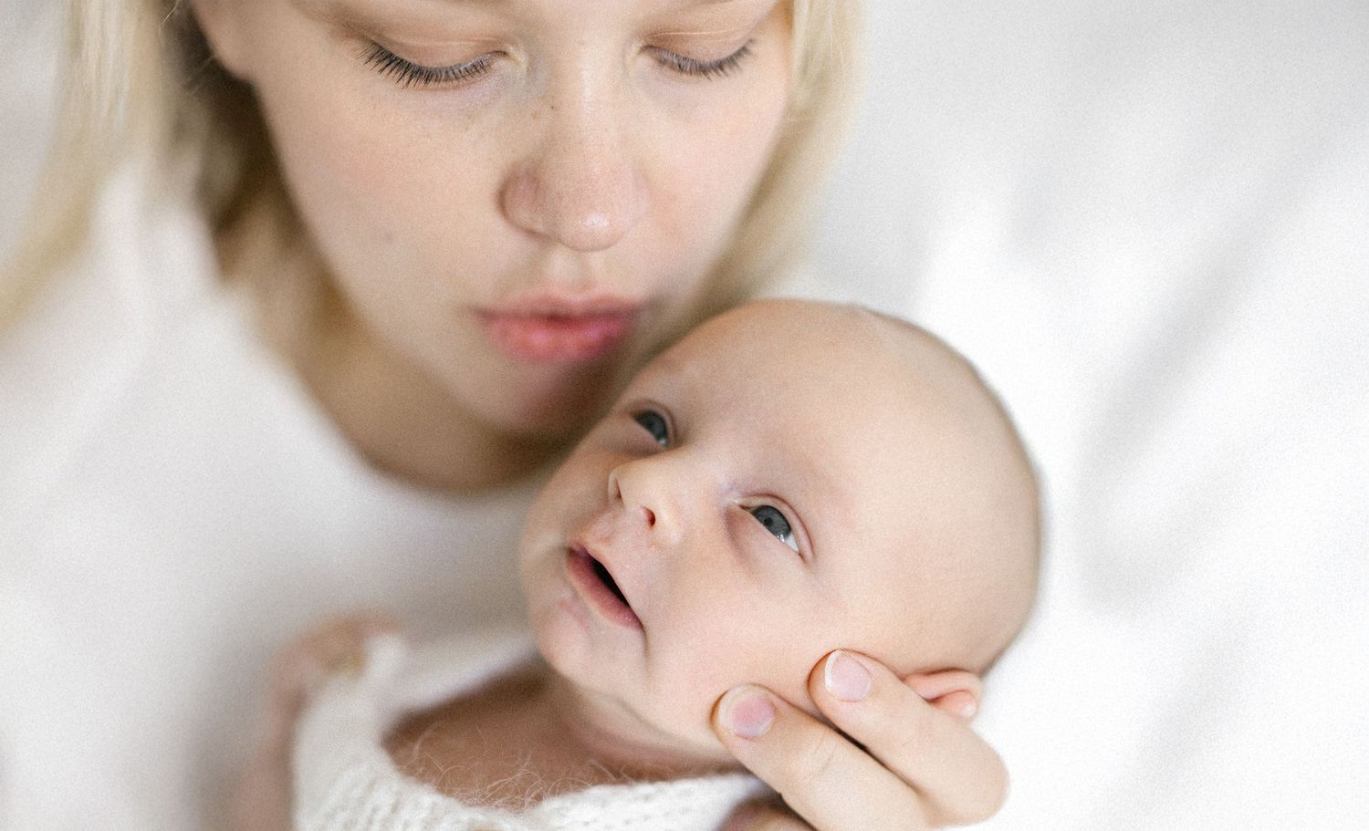 A tender moment as a blonde mother gazes lovingly at her newborn baby wrapped in a white blanket.