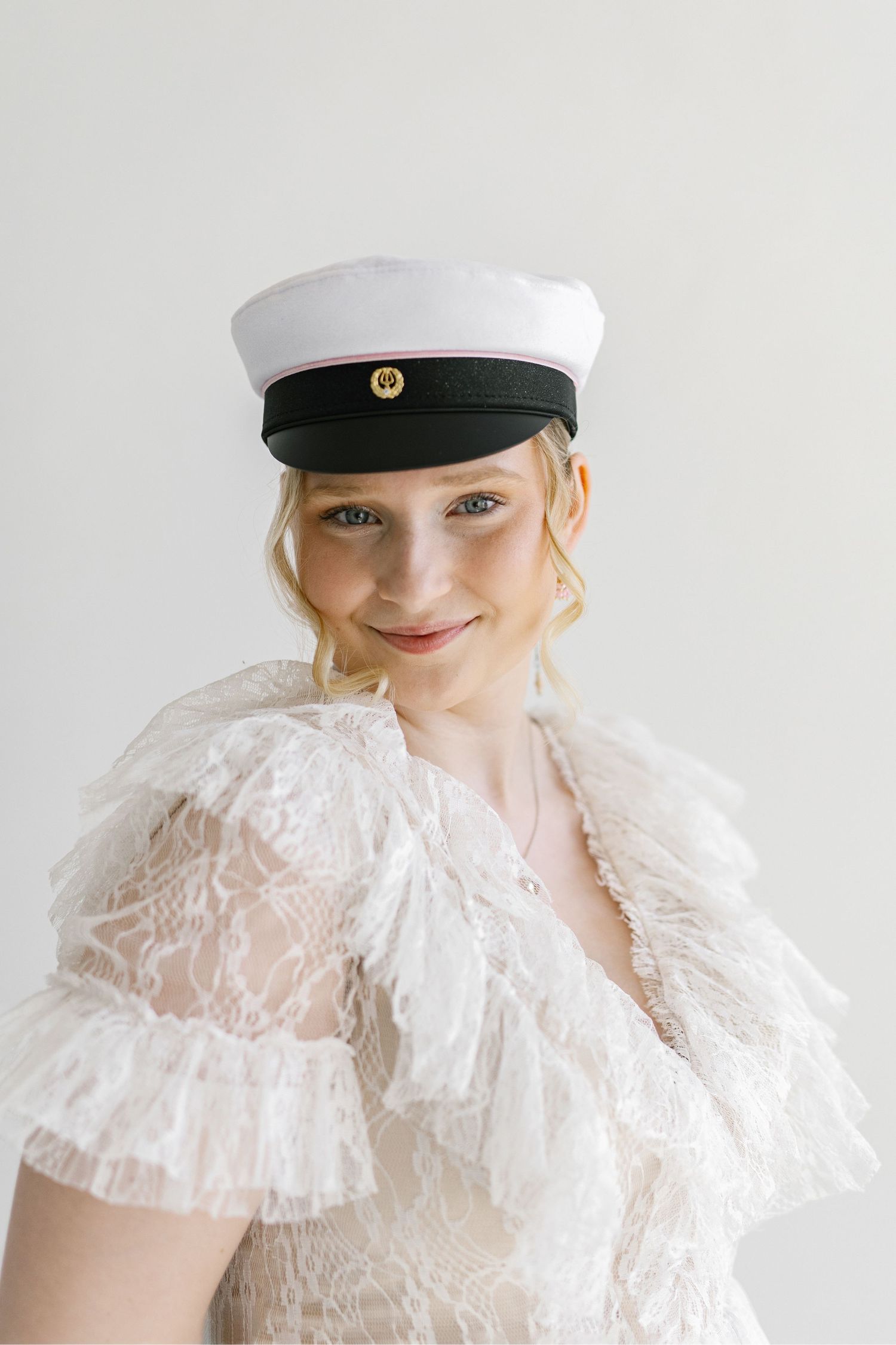 Smiling blonde woman wearing a white lace dress and white captain hat with gold emblem against a light background.