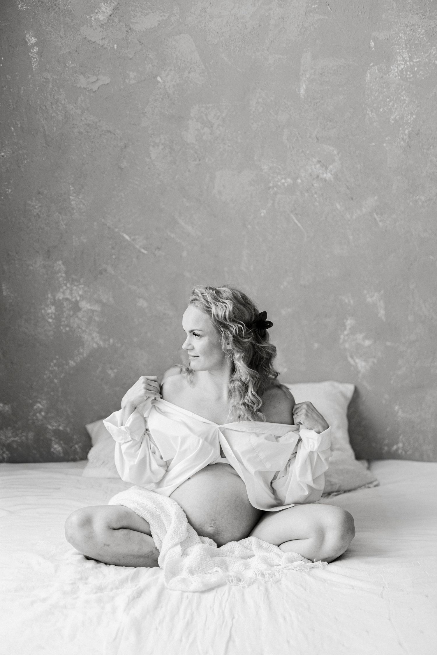 Black and white photo of a pregnant woman sitting cross-legged on a bed, opening her white shirt to reveal her baby bump.