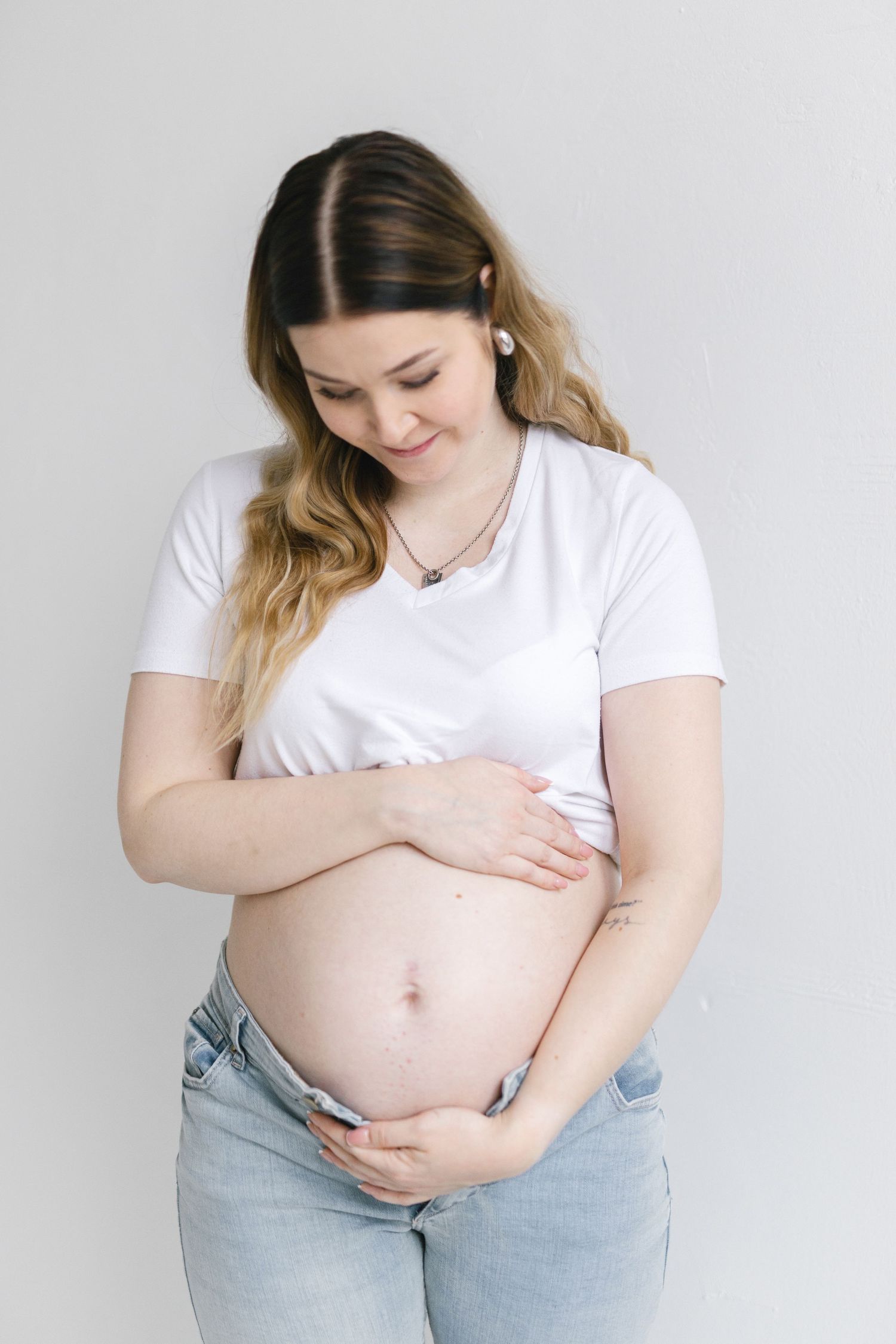 Smiling pregnant woman in white t-shirt and jeans cradling her baby bump against a light gray background.