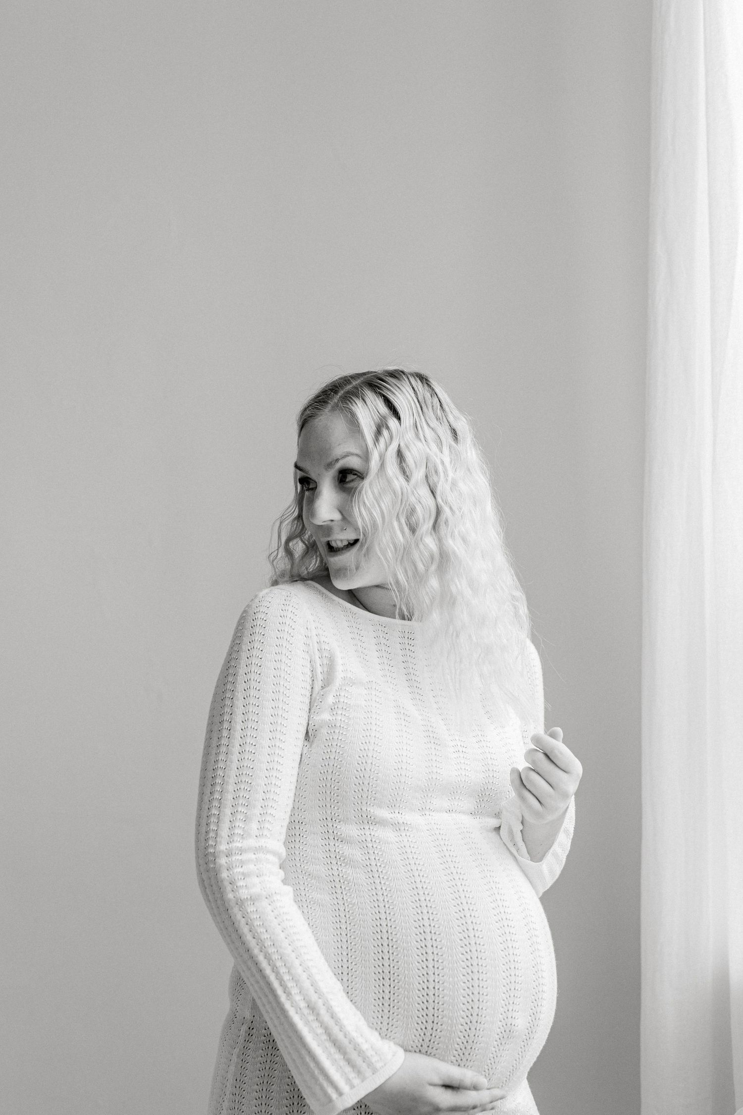 Black and white photo of a smiling pregnant woman in a white knit dress cradling her baby bump near a window.