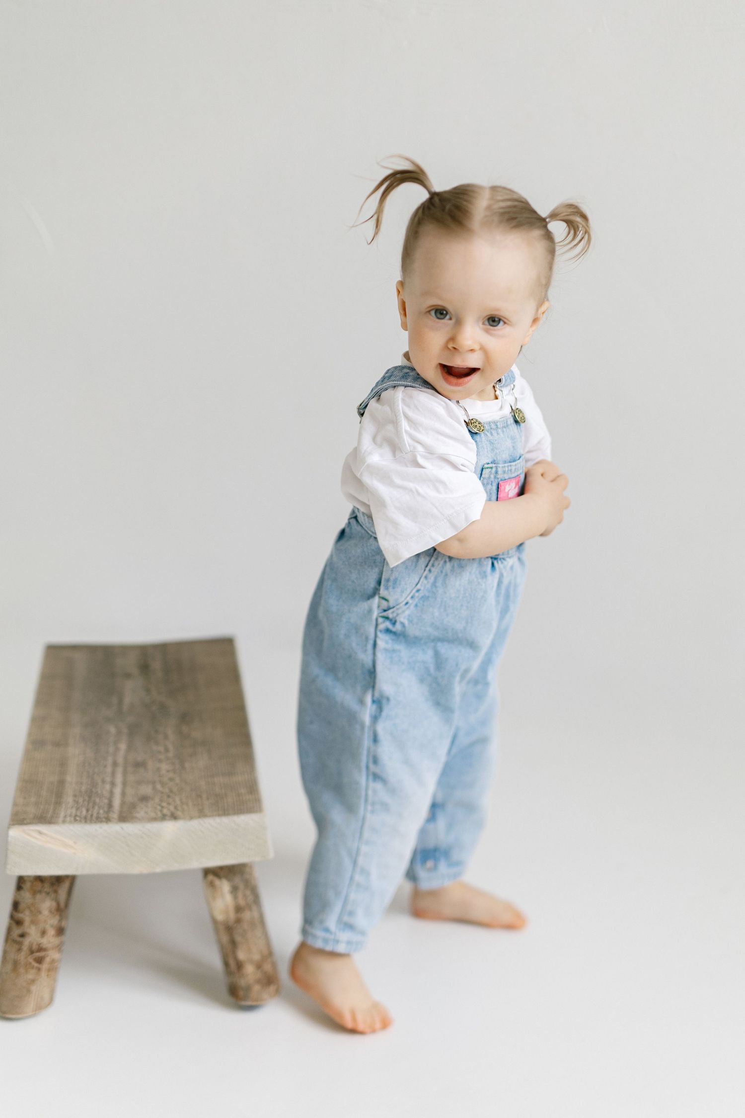 Toddler girl with pigtails wearing denim overalls and white shirt standing barefoot next to wooden bench.