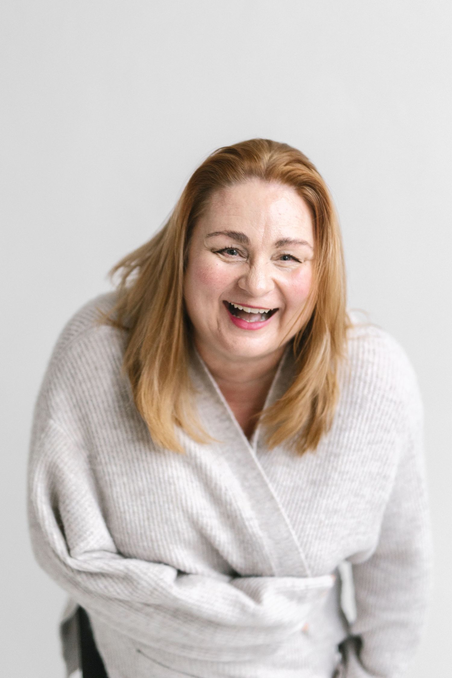 Woman wearing cozy gray sweater laughing joyfully against white background.