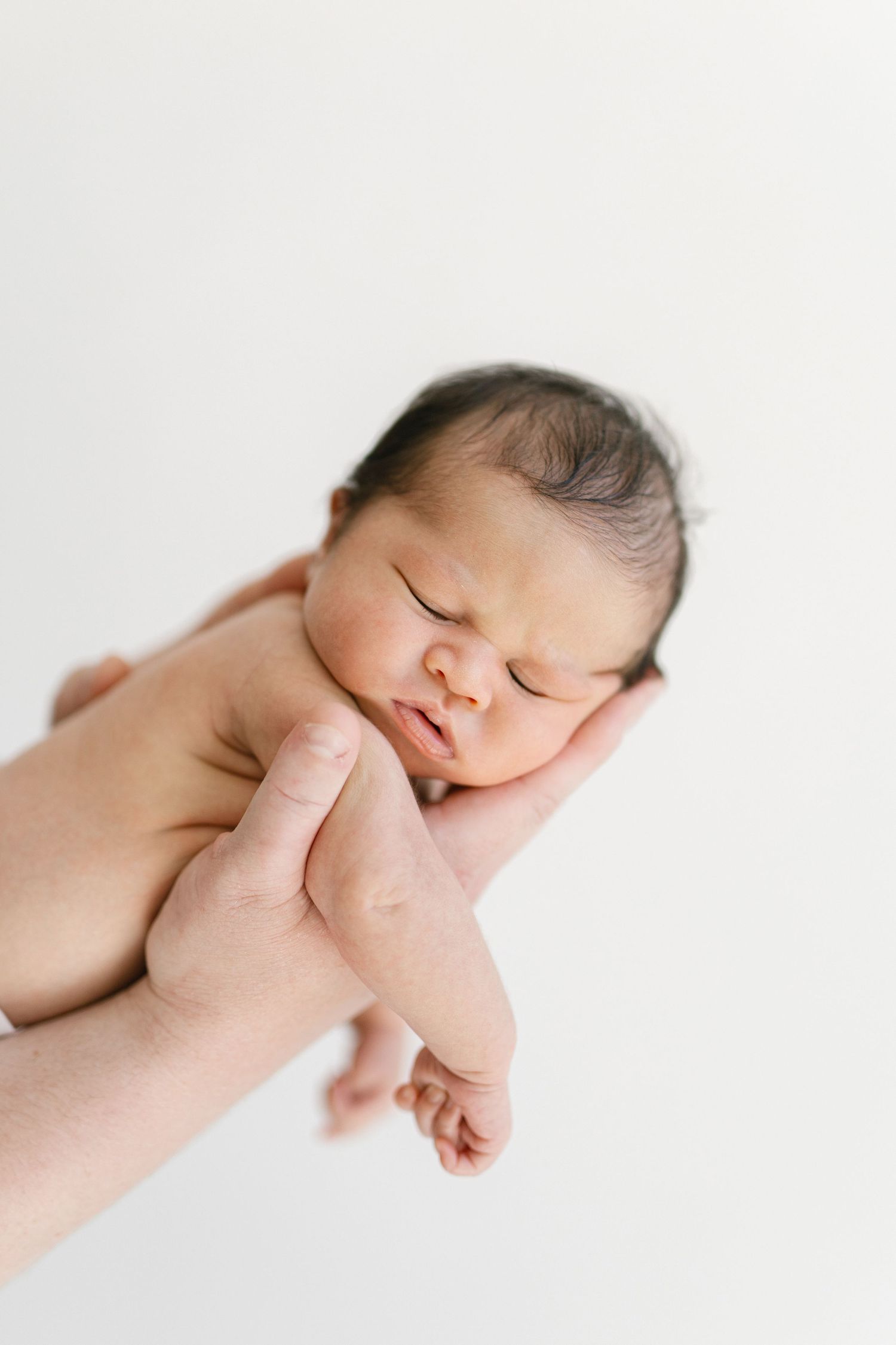 A sleeping newborn baby with dark hair rests peacefully in an adult's hands against a white background.
