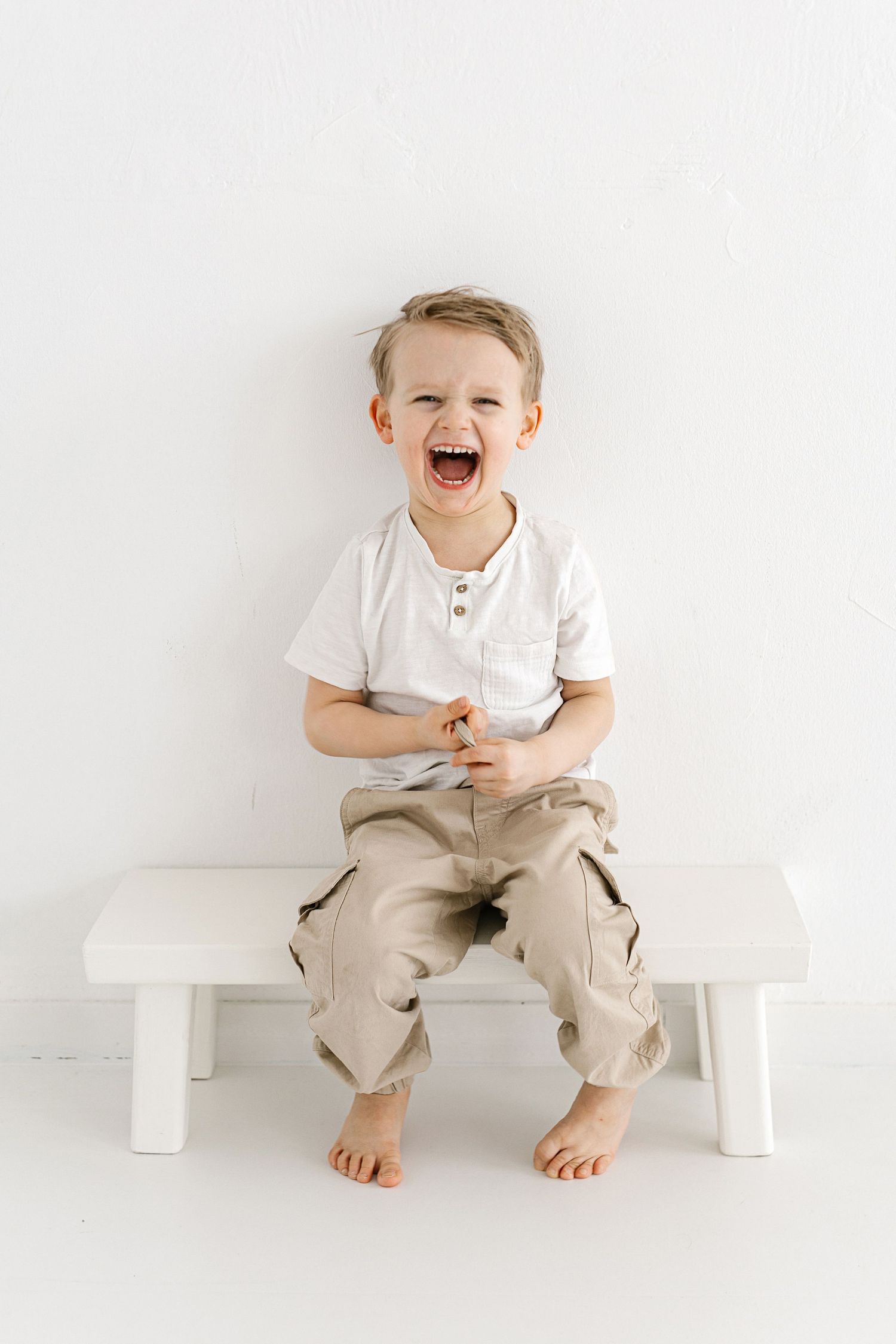 A laughing young boy sits barefoot on a white bench, wearing a white shirt and beige cargo pants.