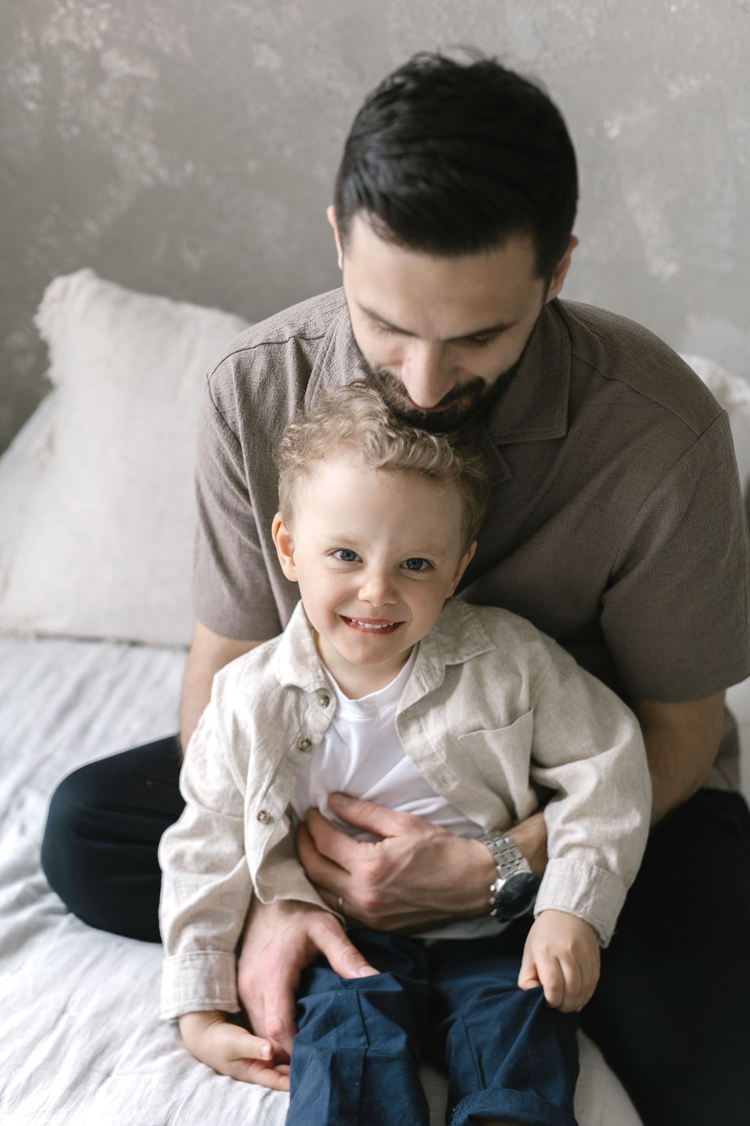 A father sits on a bed, lovingly embracing his smiling young son in matching neutral-toned outfits.