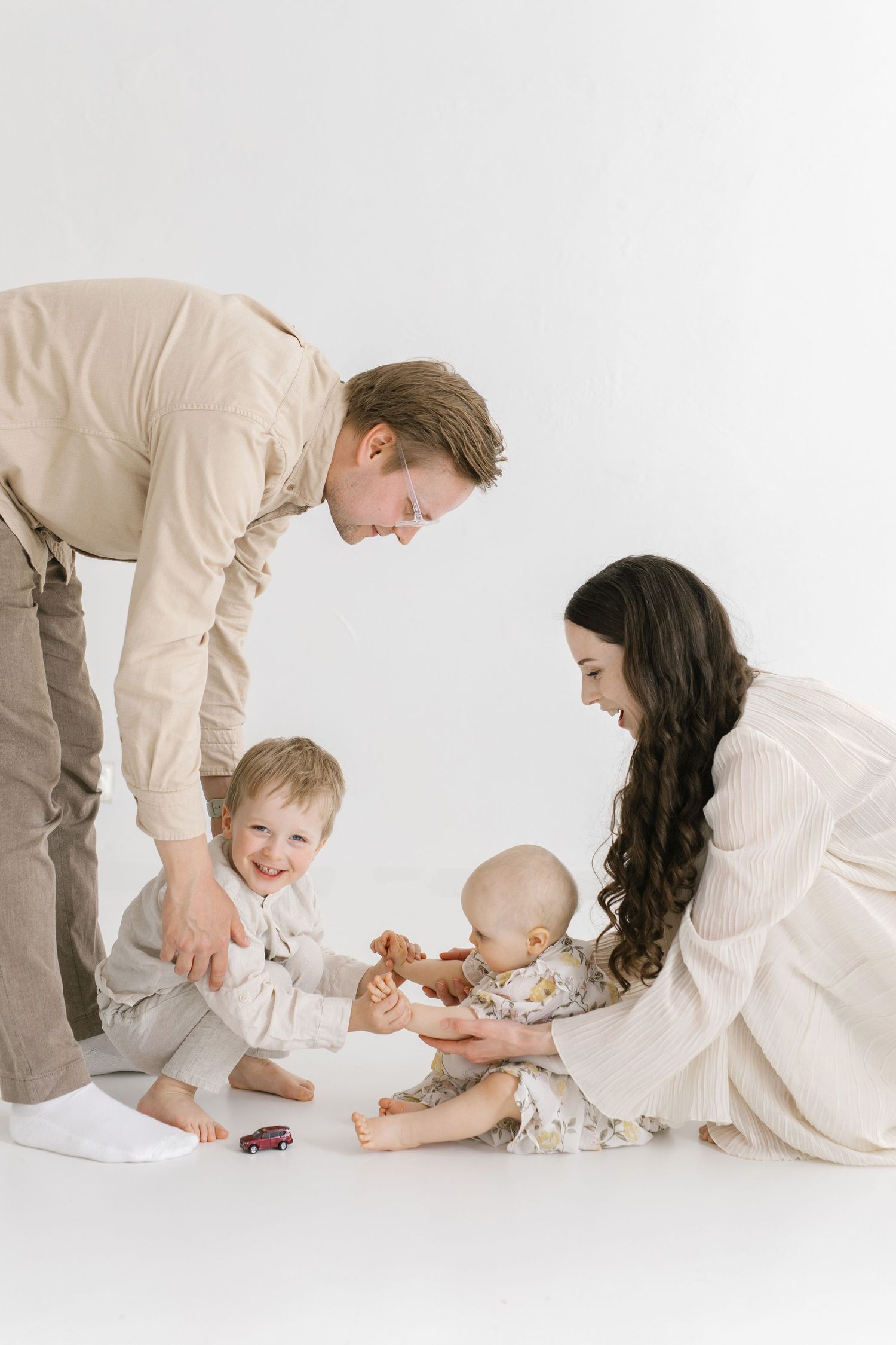 A young family with two small children playing together on a white studio floor, dressed in neutral tones.