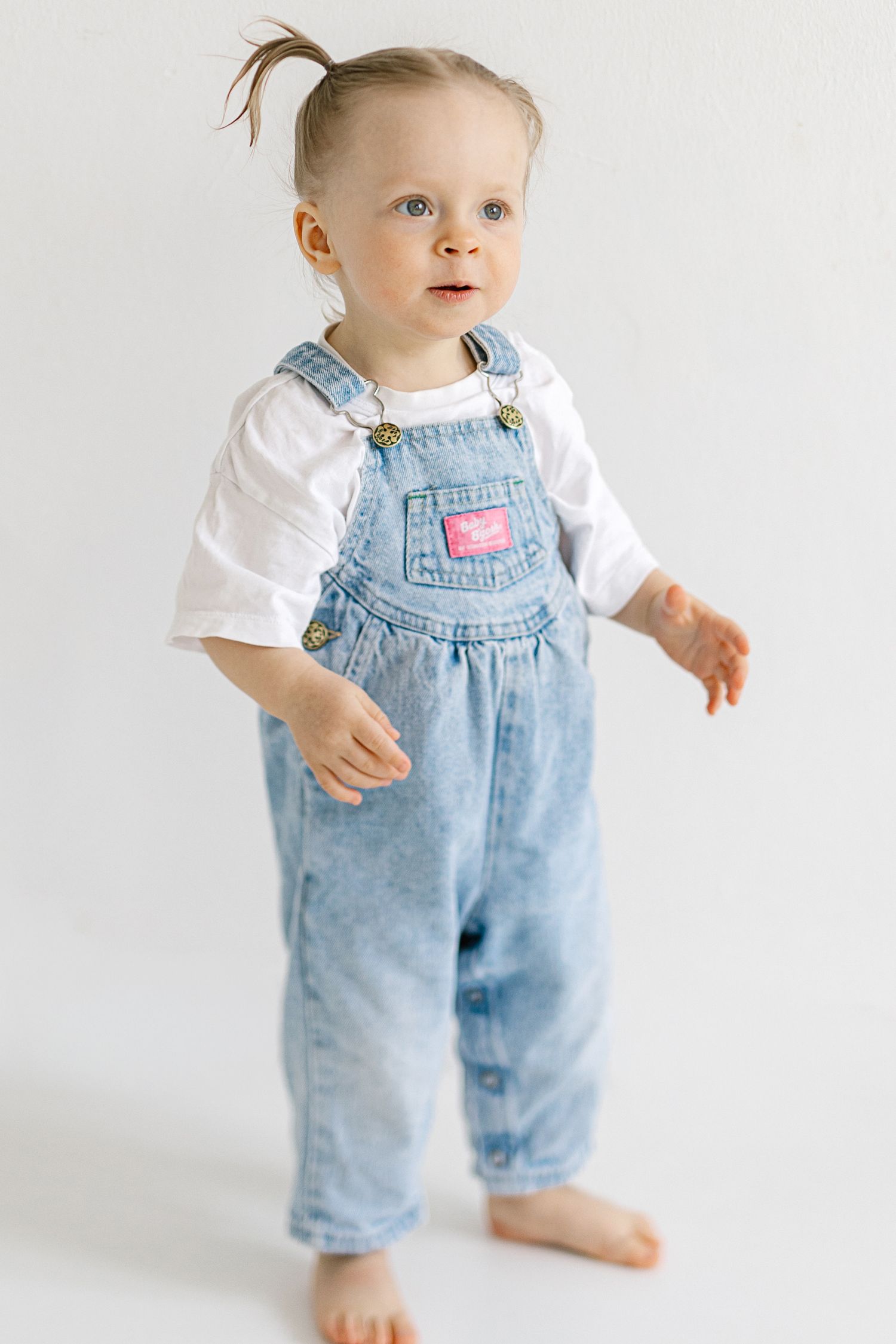 Toddler girl in denim overalls and white shirt stands barefoot against white background.